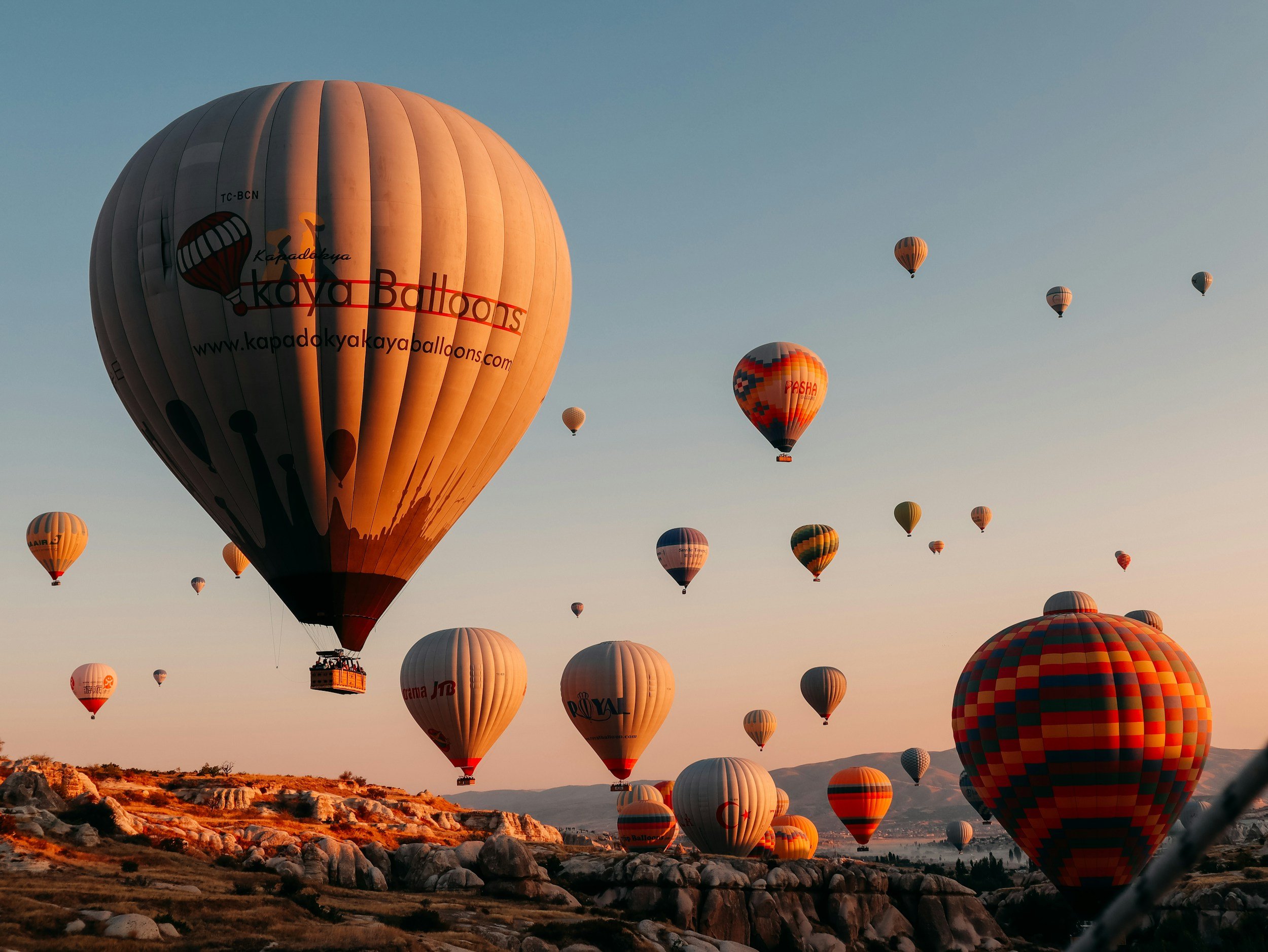 Numerous hot air balloons floating in the sky at sunrise or sunset over a rocky landscape.