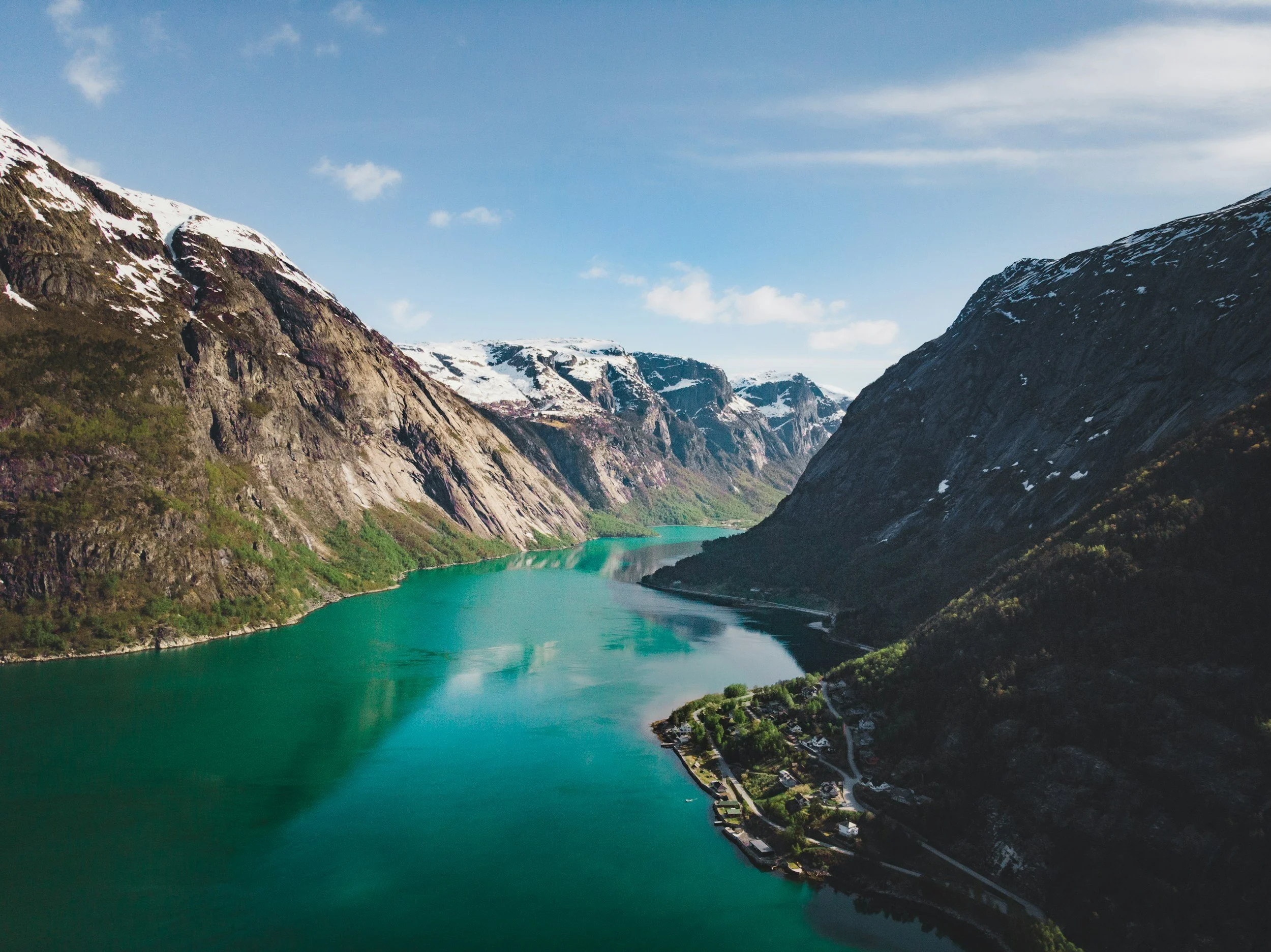 A river flowing through a mountainous valley with snow-capped peaks and a small town along the riverbank under a partly cloudy sky.