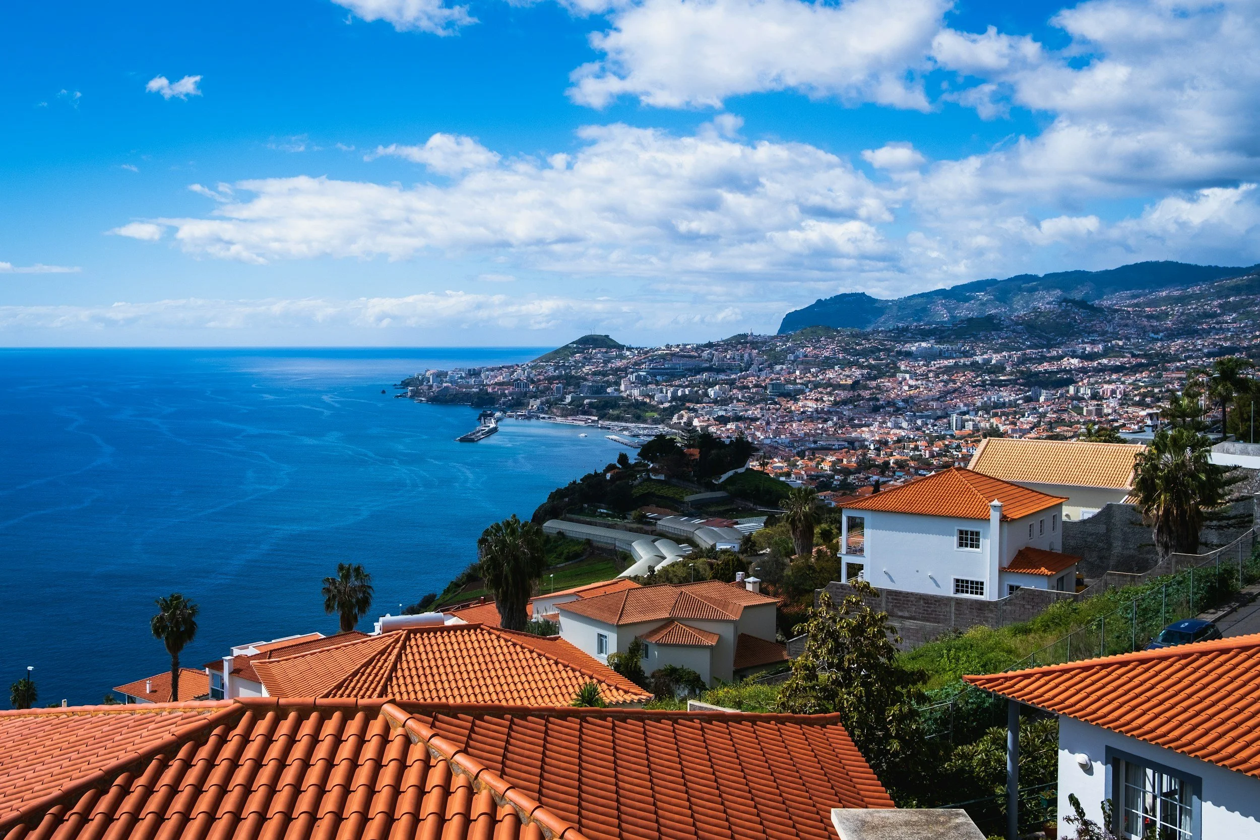 A scenic coastal town with red-tiled rooftops, overlooking the ocean under a partly cloudy sky with mountains in the background.