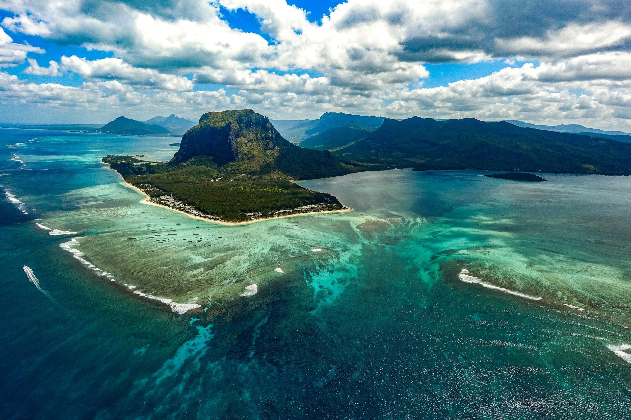 Aerial view of an island with a mountainous landscape surrounded by turquoise waters and coral reefs, under partly cloudy sky.