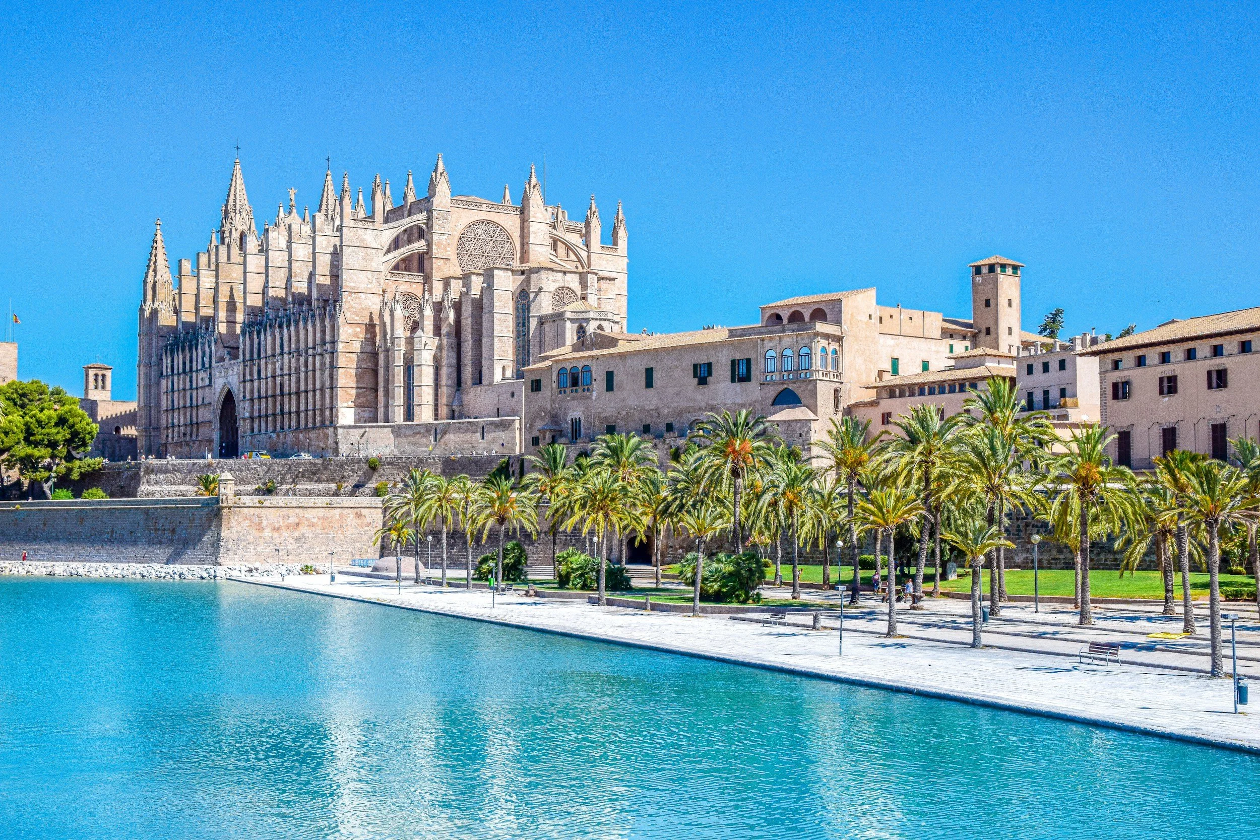 View of a historic cathedral with gothic architecture, surrounded by palm trees, next to a body of water, under a clear blue sky.