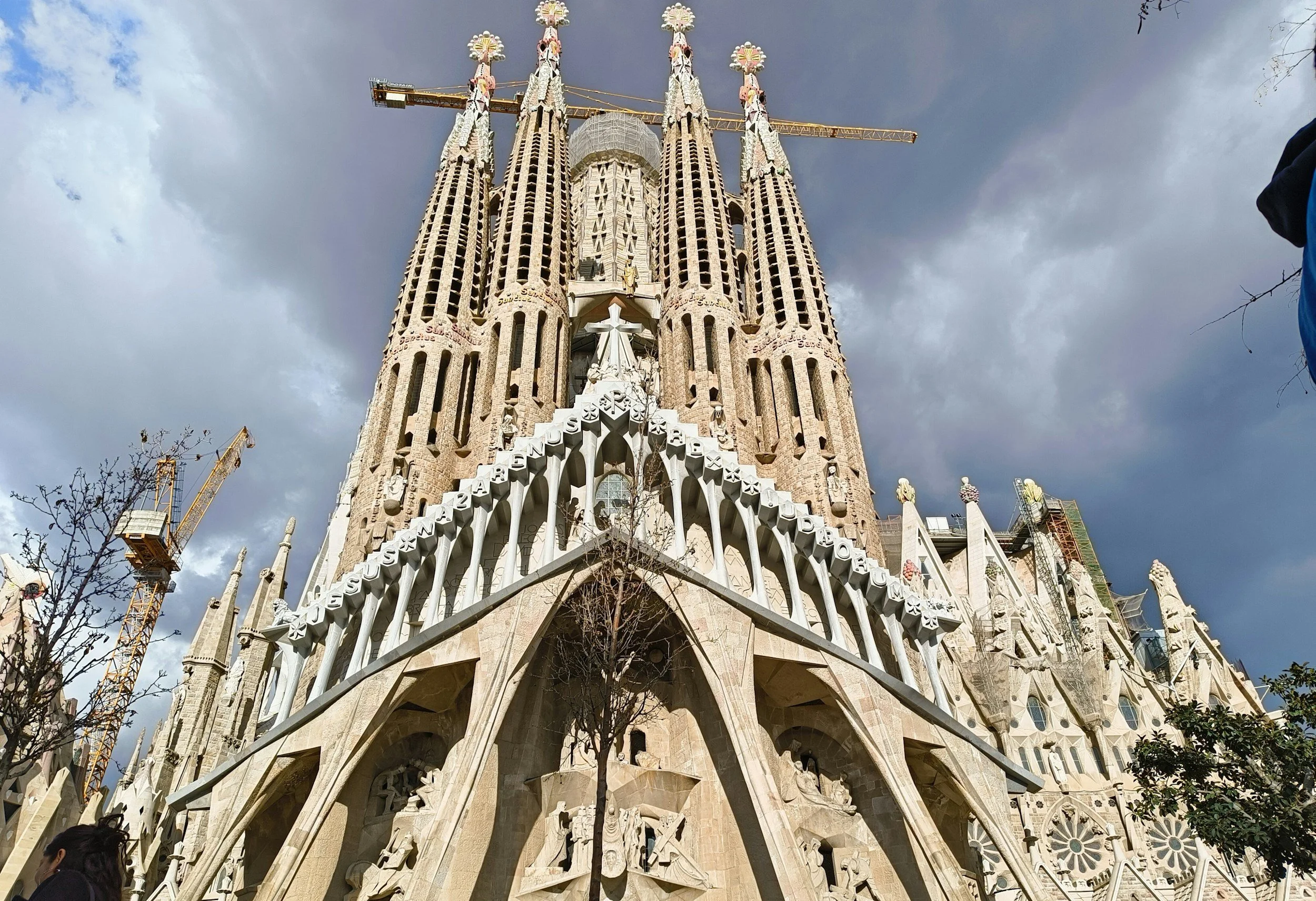 Sagrada Familia basilica in Barcelona under construction, with cranes around the towers and cloudy sky in the background.