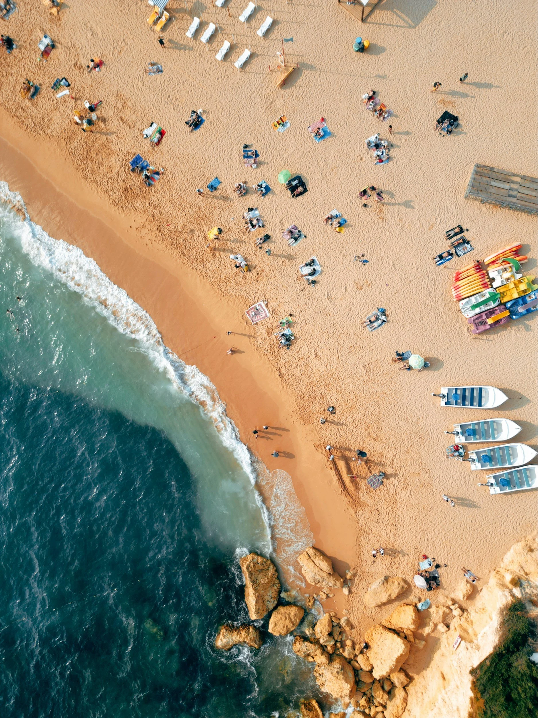 Aerial view of a sandy beach with people relaxing on chairs and towels, colorful umbrellas, and boats parked along the shoreline. The ocean water is blue-green with waves crashing onto the beach.