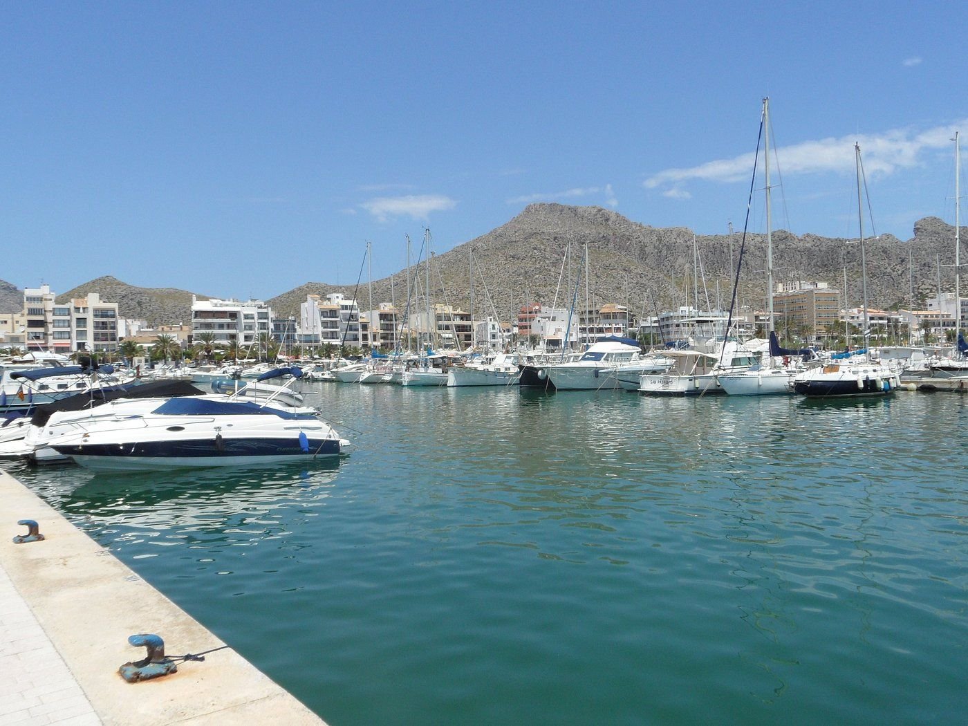 Marina with numerous sailboats and yachts docked, buildings and mountains in the background under a clear blue sky.