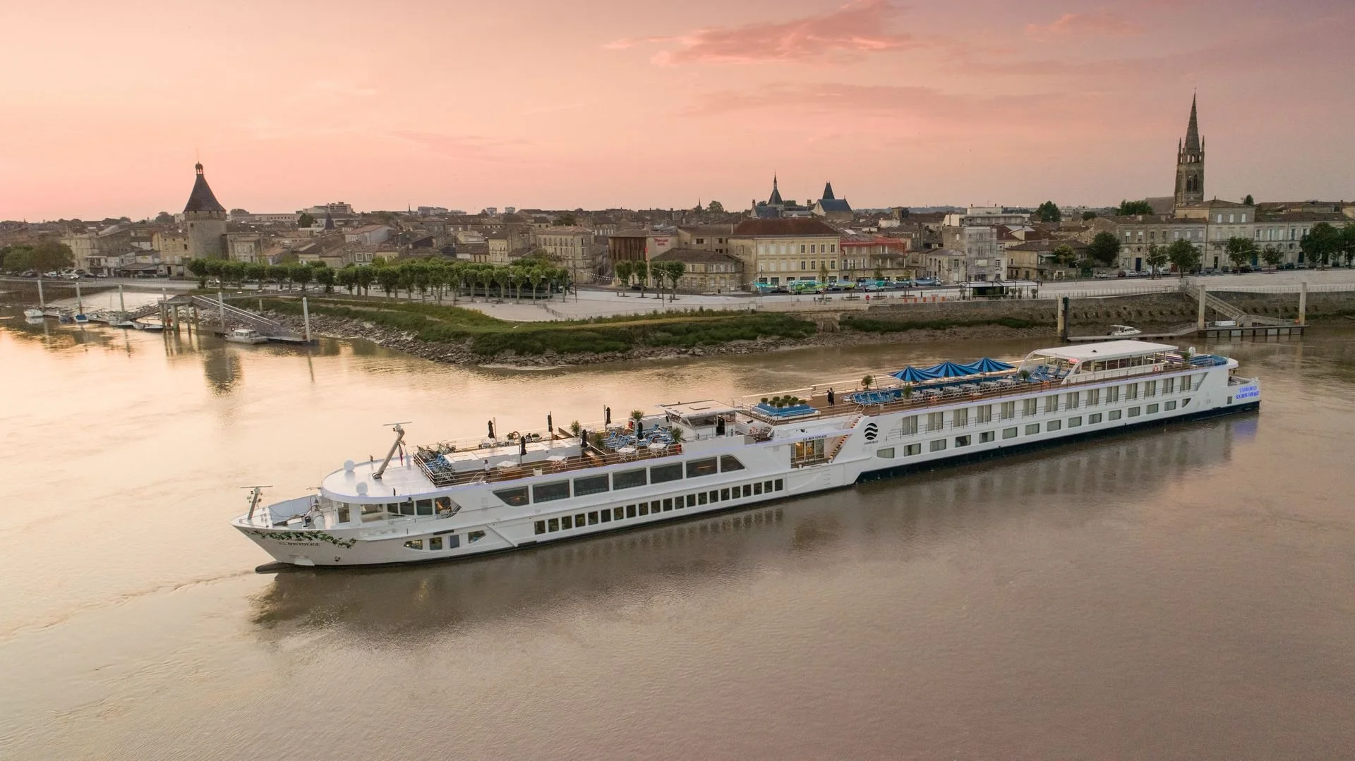 A large white cruise ship on a river with a town and historic buildings in the background at sunset.