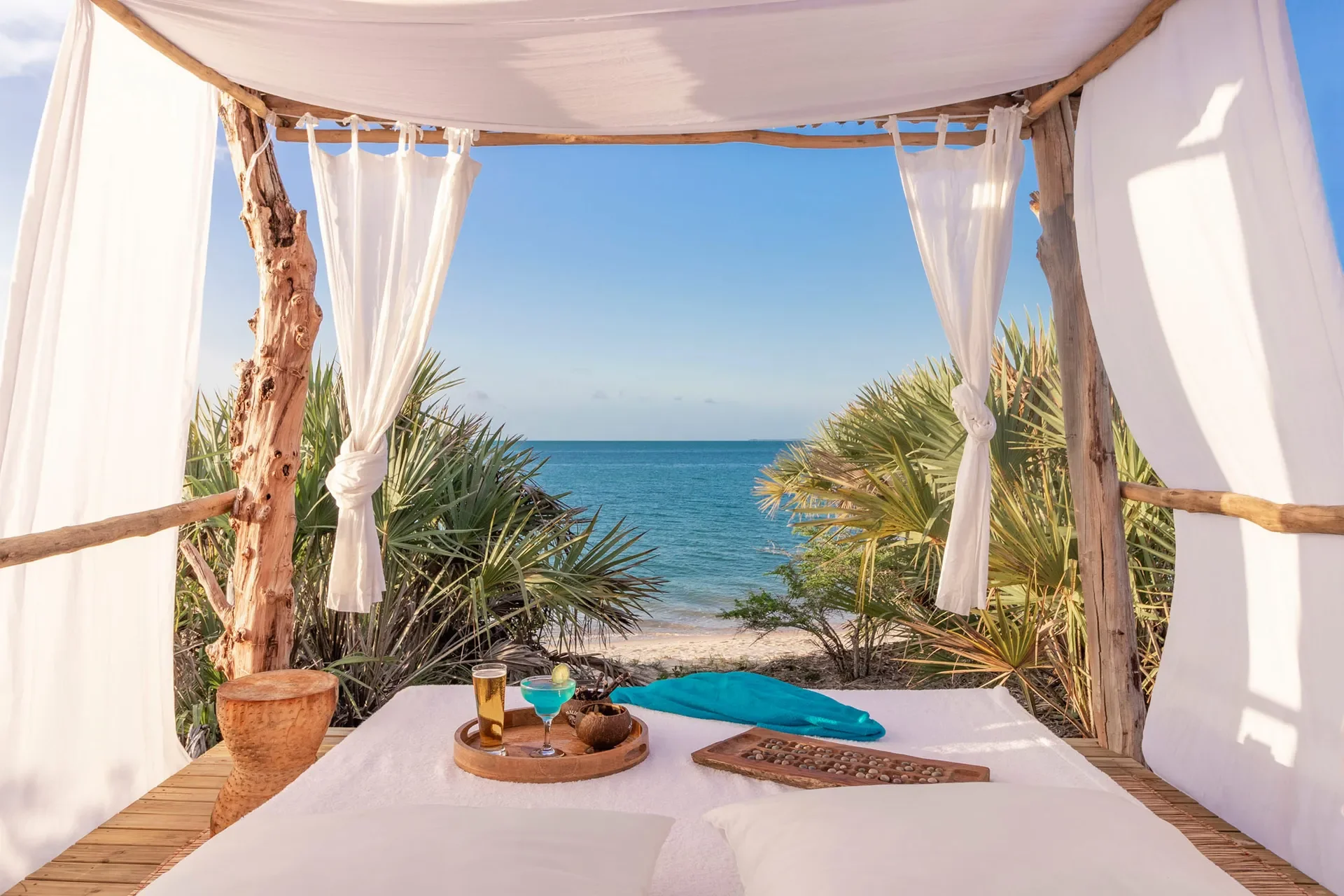 Beach cabana with white curtains and rooftop view of the ocean, with drinks and snacks on a table, surrounded by tropical plants.