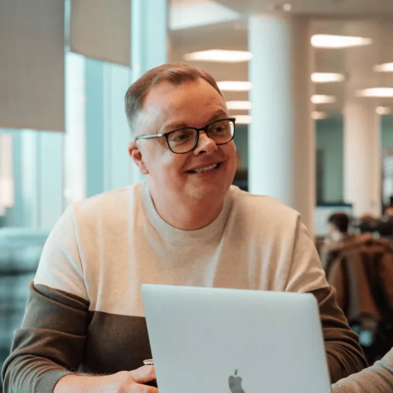 A smiling man with glasses sitting at a table with a silver laptop in a modern, well-lit space.