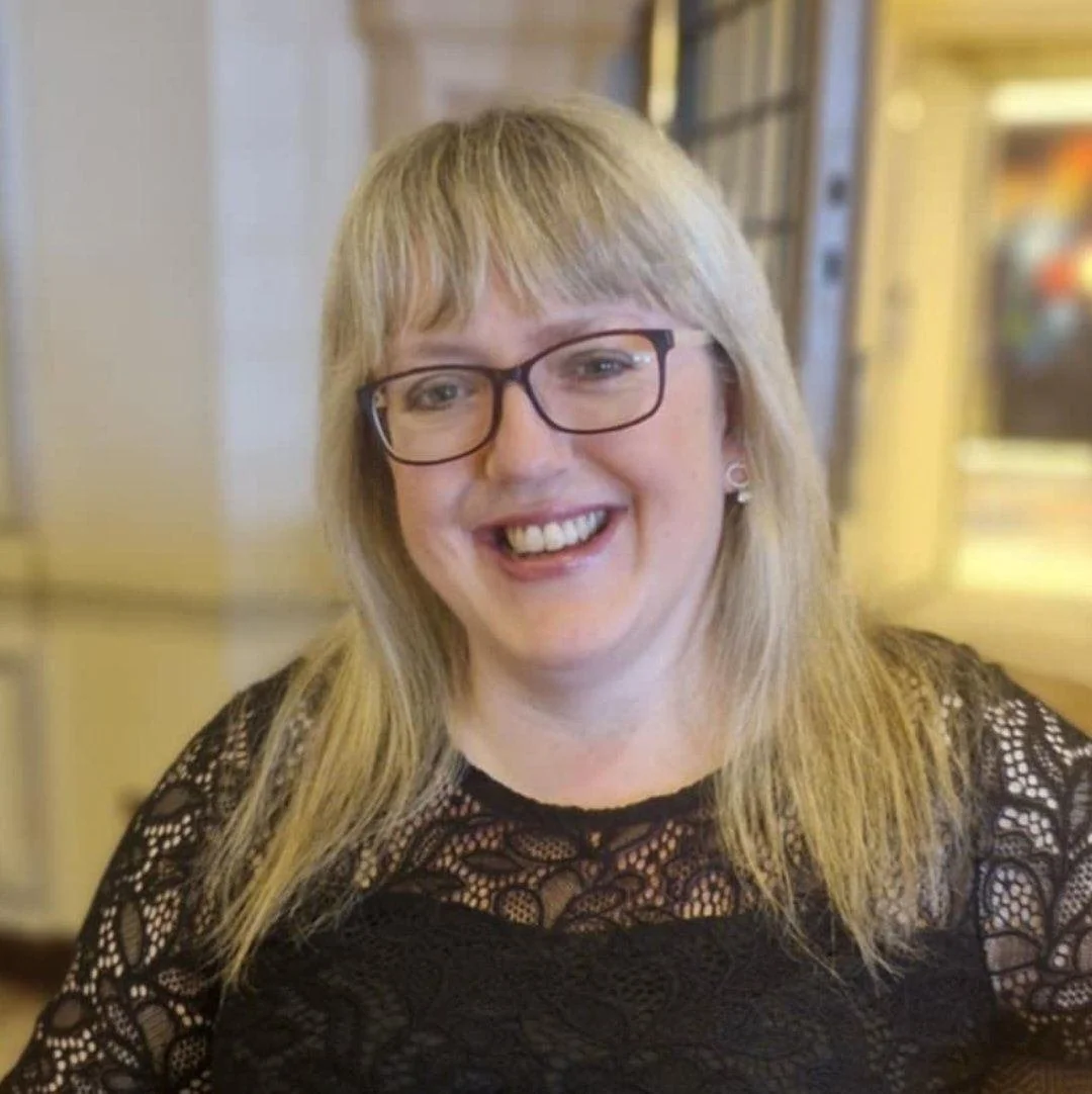 A woman with blonde hair, glasses, and a black lace top, smiling in a restaurant or cafe.