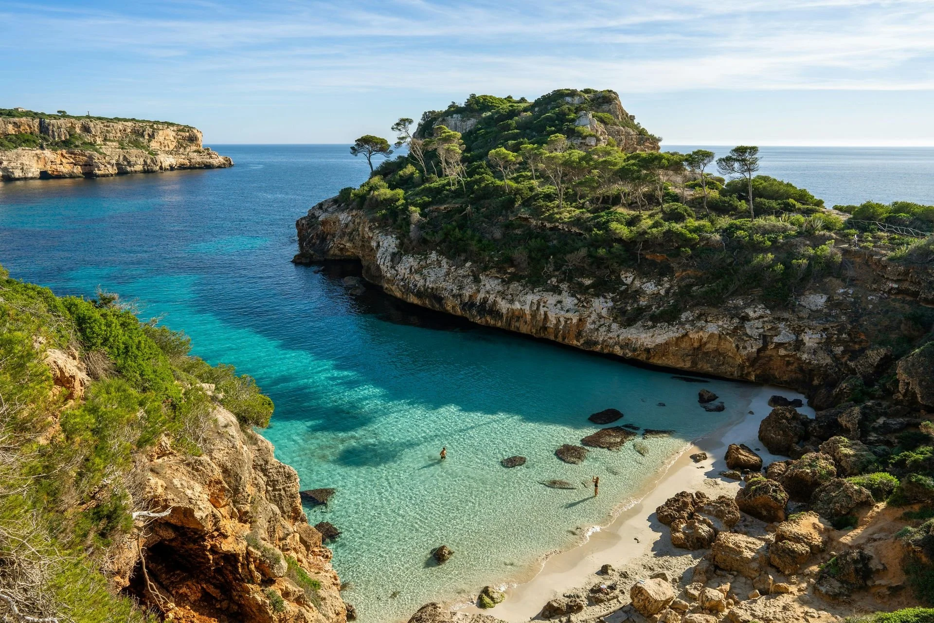A secluded beach with turquoise water, surrounded by rocky cliffs and lush greenery, with a few people enjoying the sand and water under a partly cloudy sky.