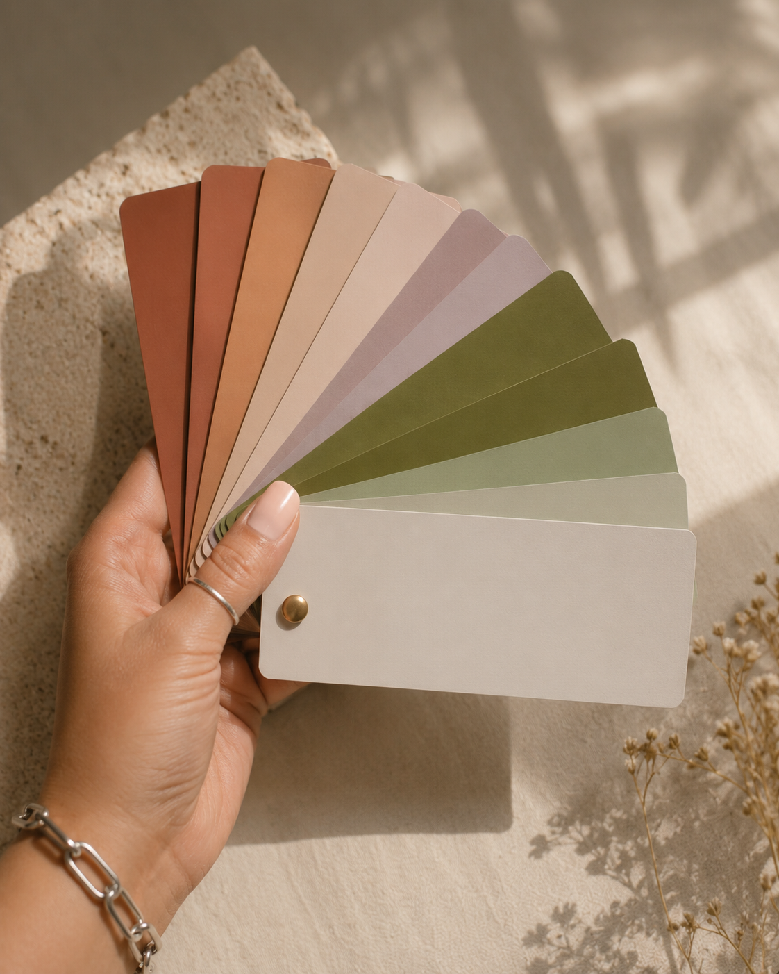 Hand holding a fan of color swatches in earthy tones, with a white blank card at the bottom, against a textured concrete surface and dried flowers.