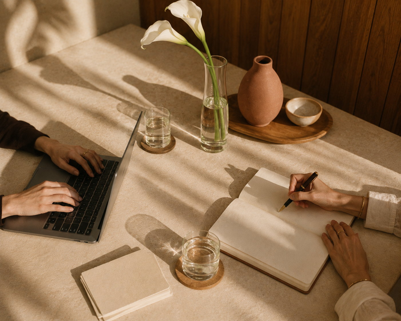 A table with a person working on a laptop and another person writing in a notebook, with a vase of white flowers, glasses of water, and decorative pottery on the table.