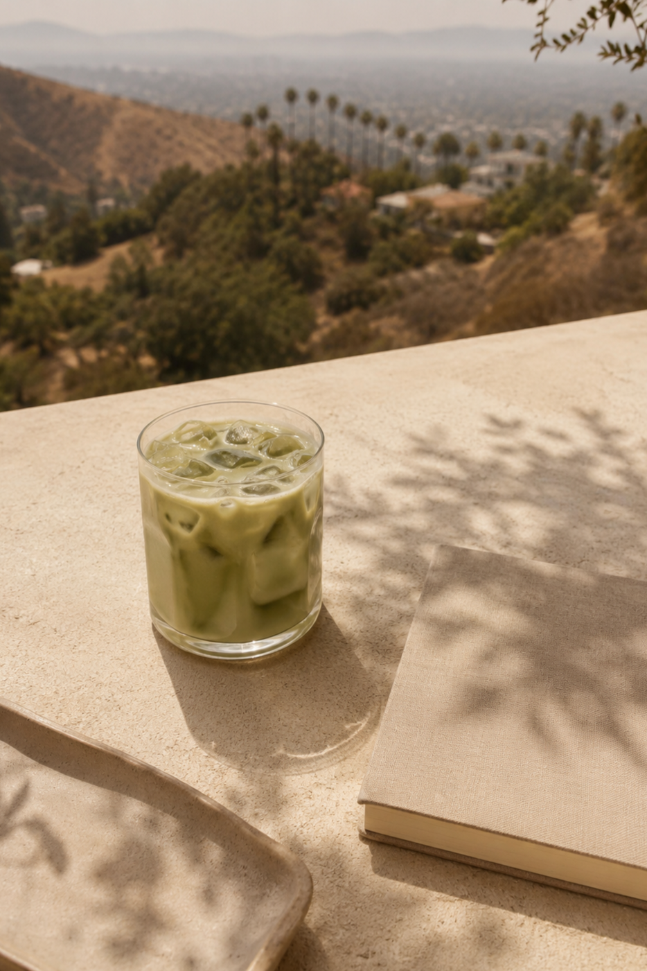 A glass of iced matcha latte on a beige outdoor table with a book and a small tray, overlooking a hilly landscape with trees and distant city view, under a partly shaded environment.