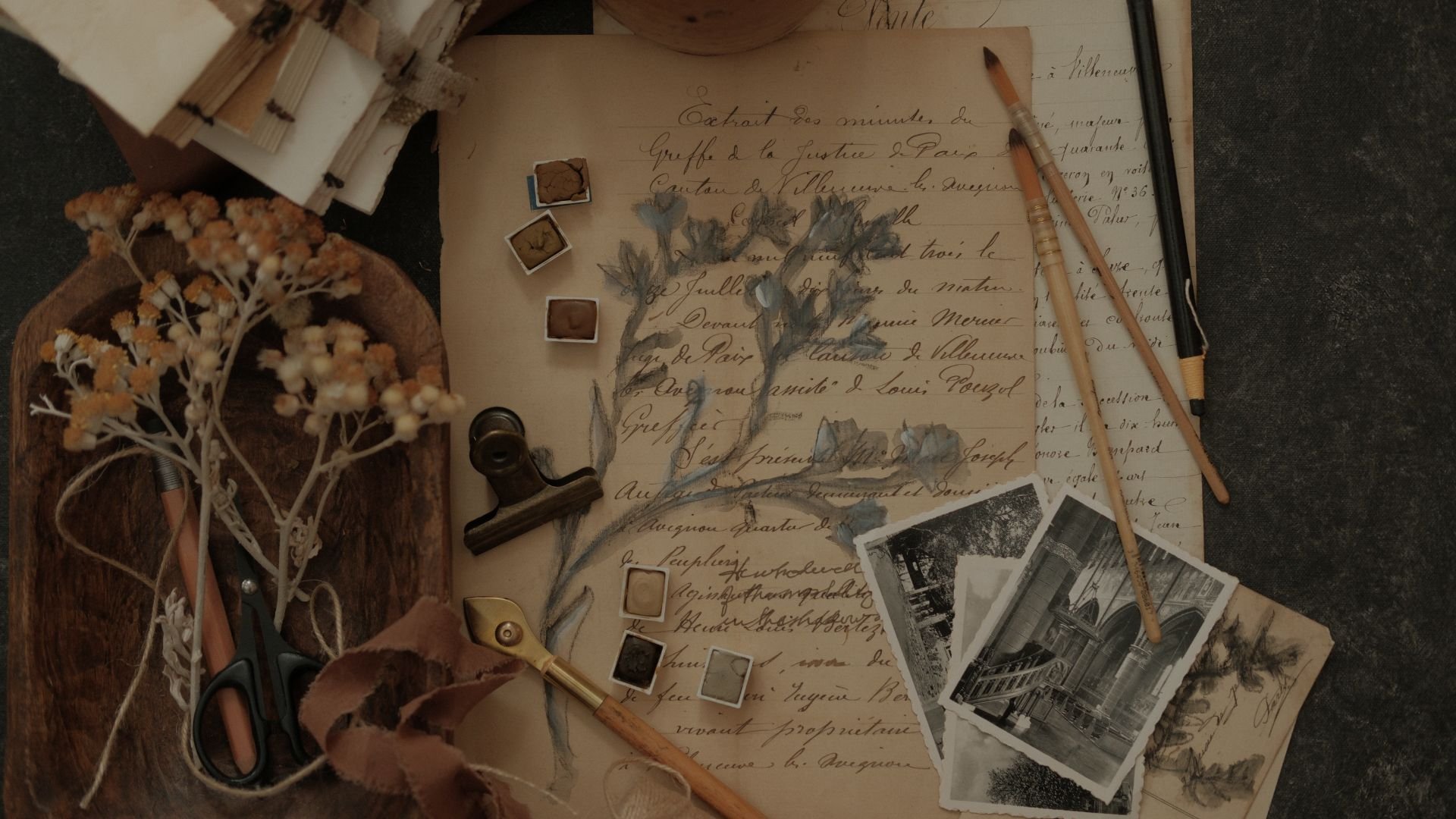 A vintage desk scene with handwritten letter, black-and-white photographs of architectural interiors, dried flowers, watercolor sketches of flowers, pencils, scissors, and small ink pads.