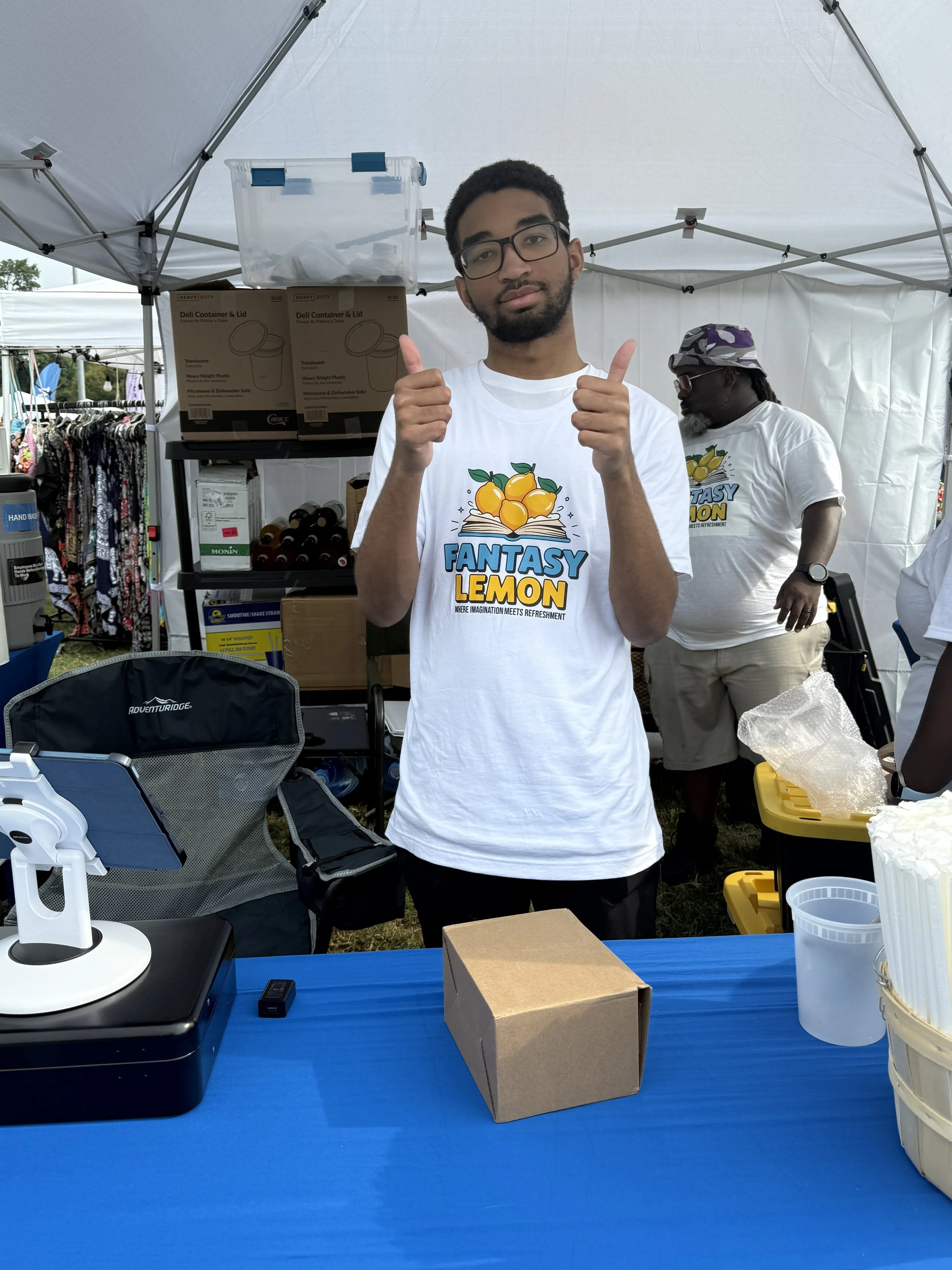 A young man wearing glasses and a white T-shirt with a lemon and book graphic, giving a thumbs-up with both hands, standing behind a blue table at an outdoor event under a white canopy.