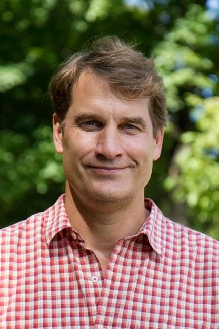 A man with light brown hair smiling outdoors, wearing a red plaid shirt, with green trees in the background.
