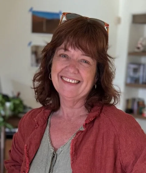 A woman with auburn hair smiling, wearing a red jacket and sunglasses on her head, indoors with a blurred background including pictures and plants.