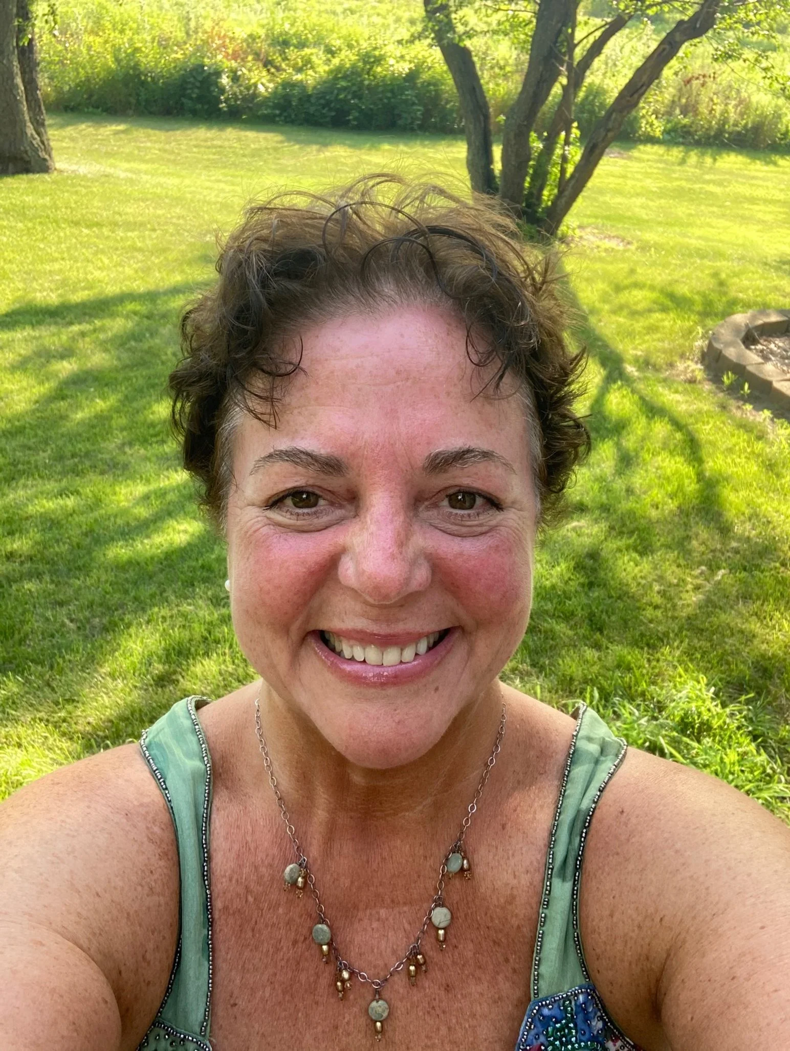 A smiling woman taking a selfie outdoors in a grassy area with trees in the background, wearing a colorful sleeveless top and a necklace.