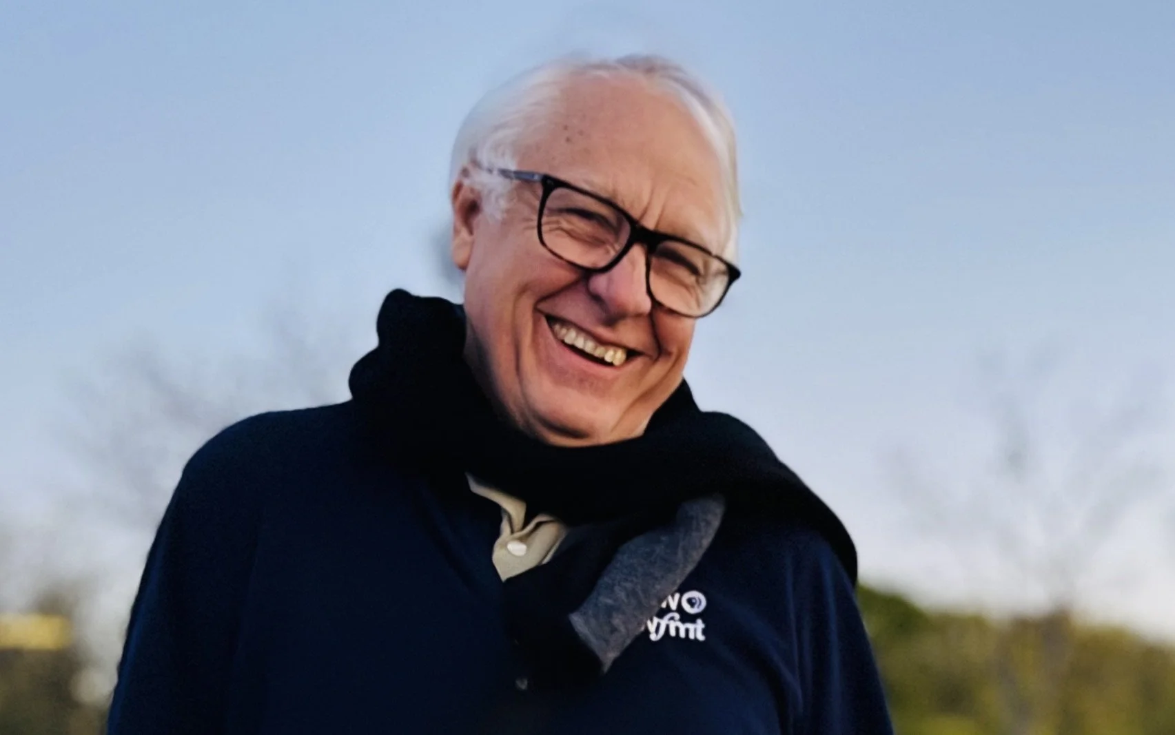 An elderly man with white hair, glasses, and a wide smile, wearing a dark jacket with a logo, outdoors during daytime.