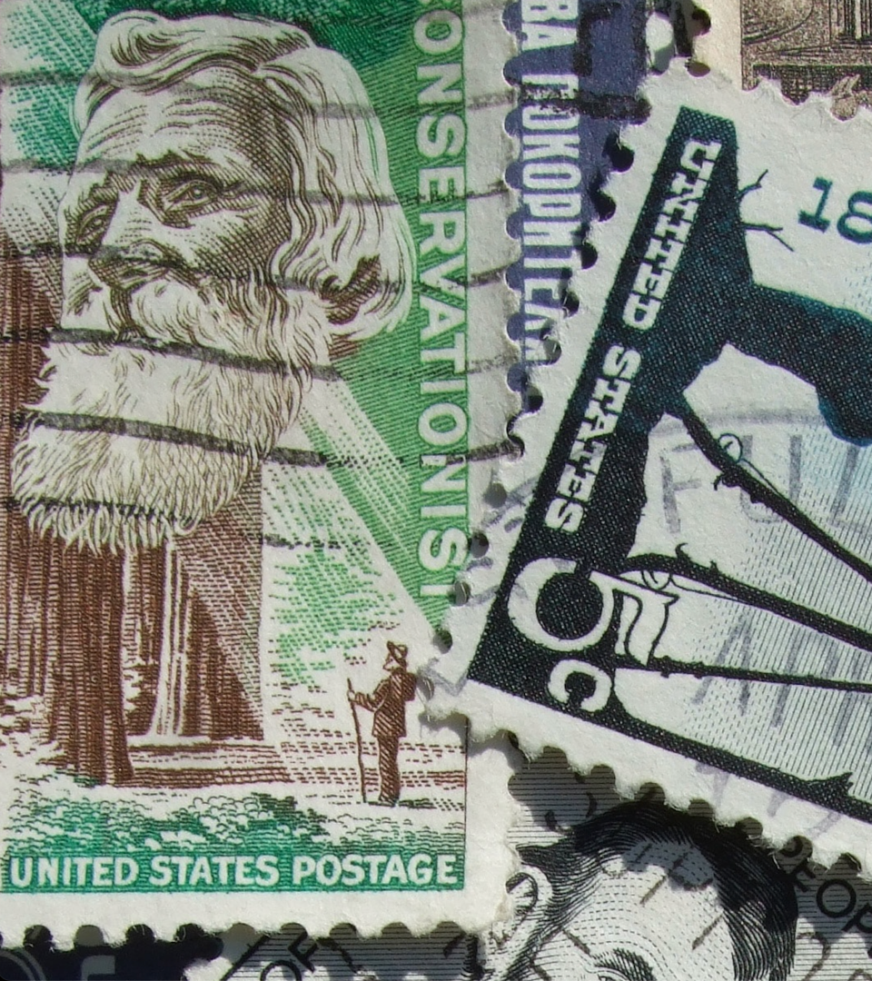 Close-up of overlapping U.S. postage stamps, featuring an illustration of a bearded man with flowing hair and a figure holding a flag.