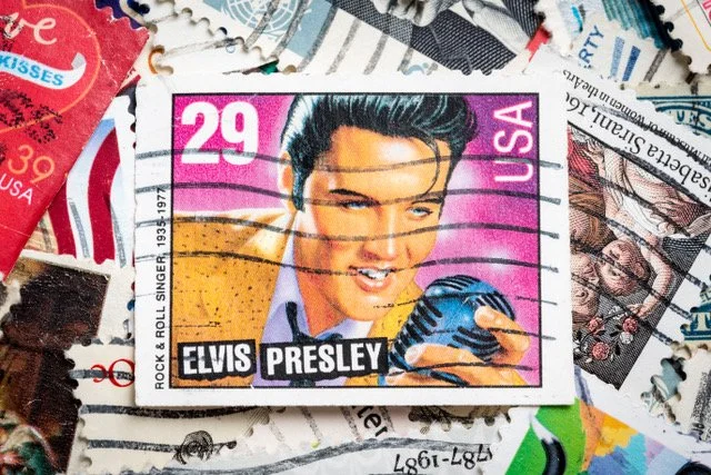 A vintage Elvis Presley postage stamp featuring a portrait of Elvis with styled hair, holding a microphone, with a pink background and the denomination 29 cents.