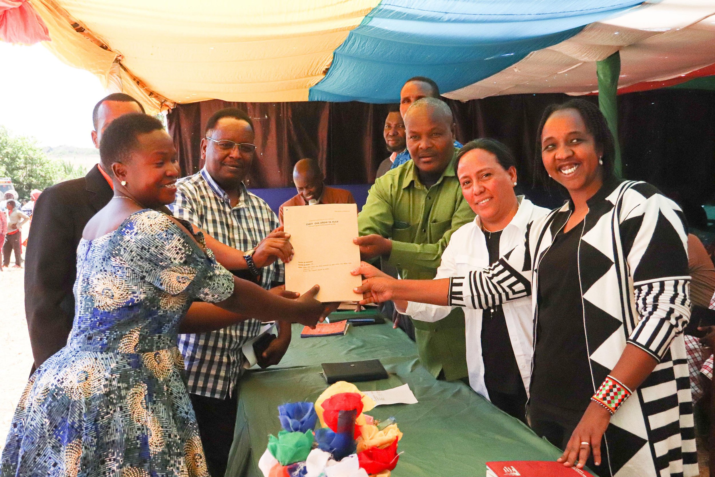 A group of people at an outdoor event, with five individuals in focus, exchanging documents, with a table decorated with colorful paper flowers in front of them, under a canopy with multicolored fabric in Northern Tanzania.