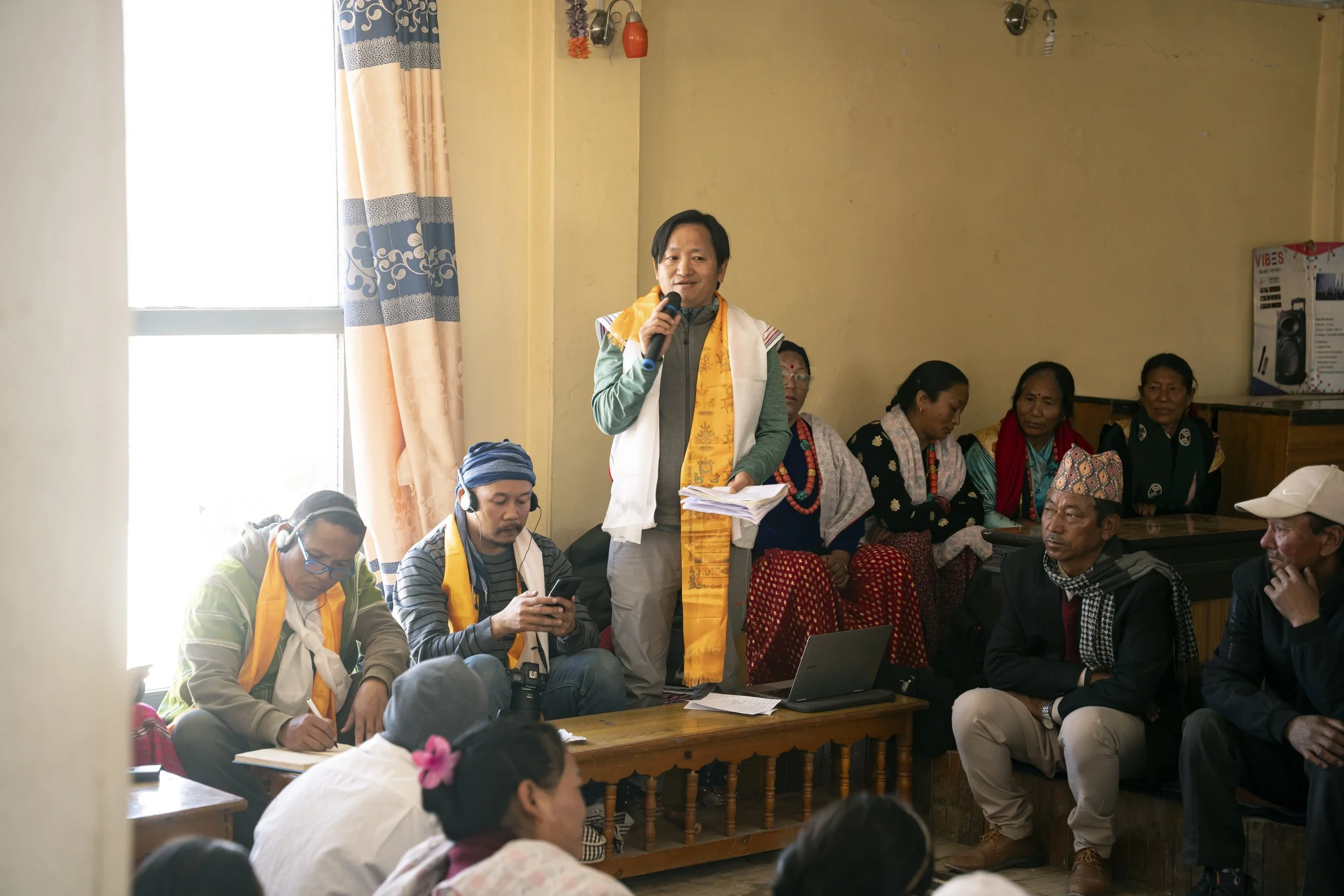 An Indigenous Nepalese man standing and speaking into a microphone at a community gathering, surrounded by seated people including women in traditional clothing and men, in a room with natural light, beige walls, and a laptop on the table in front.