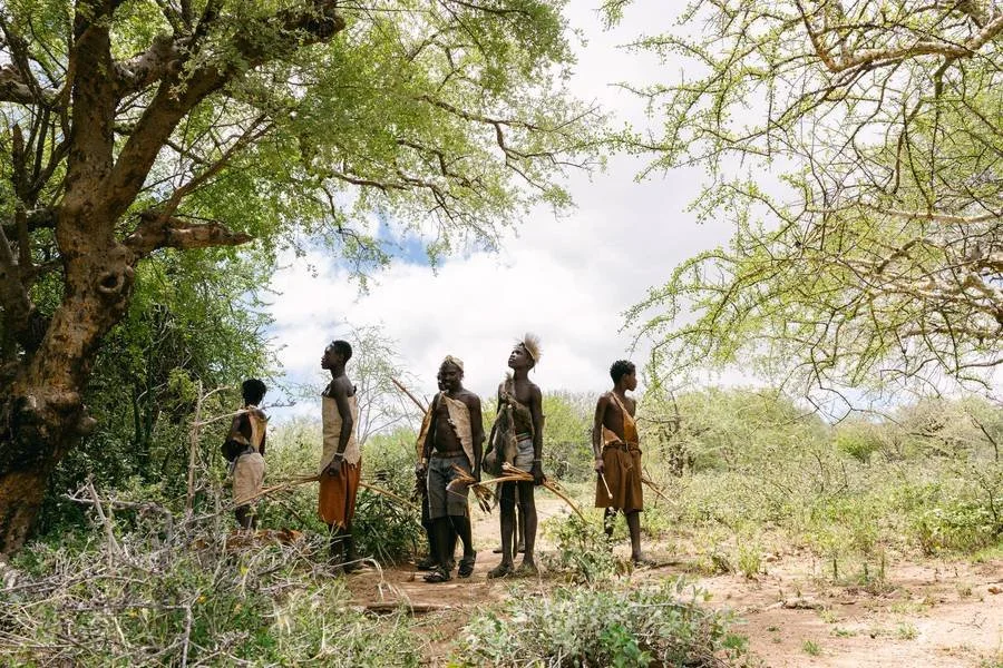 Group of five Indigenous young children, some holding bows and arrows, standing in a semi-arid landscape with sparse vegetation and large trees during daytime in Northern Tanzania.