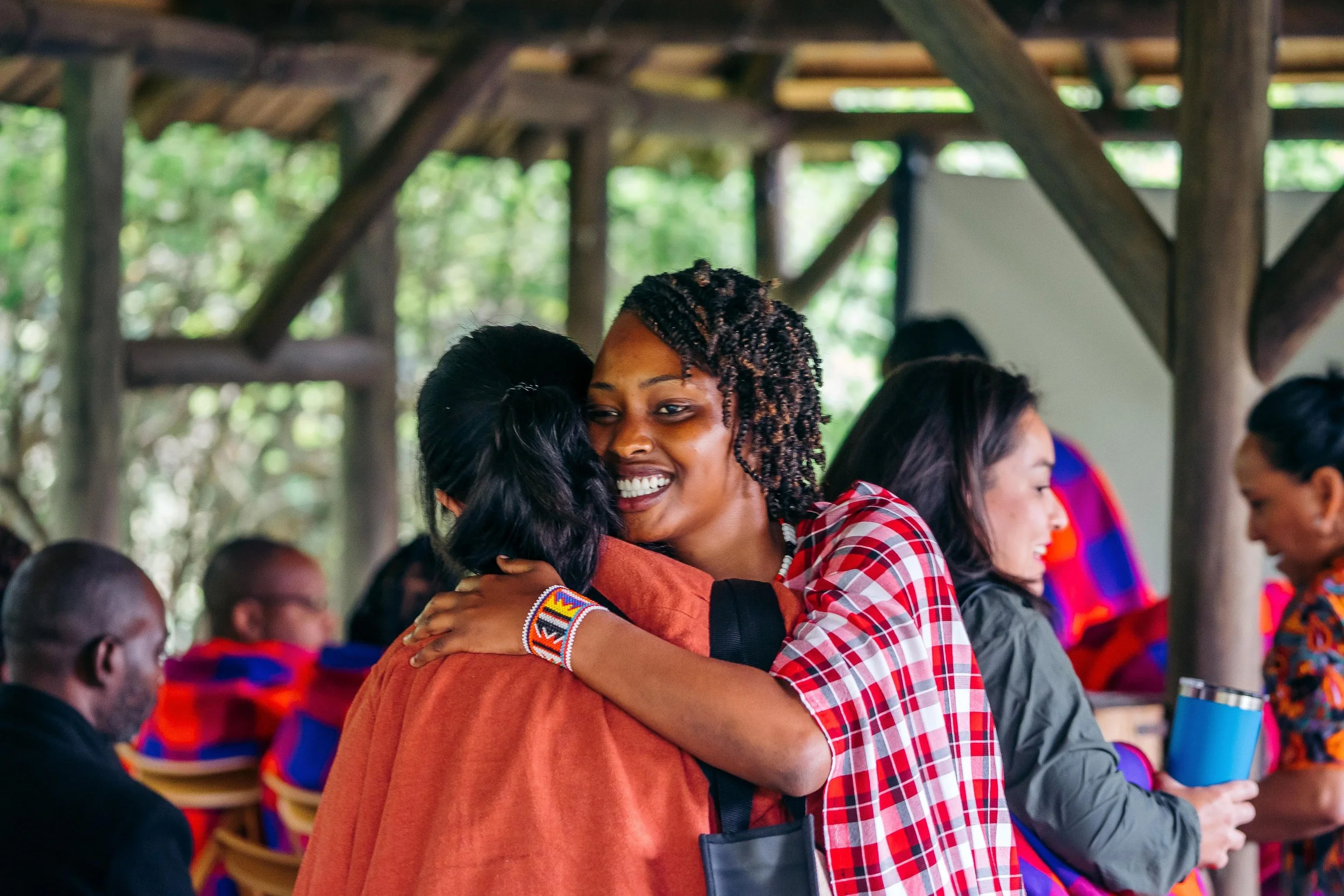 Two women hugging warmly at a gathering, with other people in the background. The setting appears to be a rustic outdoor or semi-outdoor space.