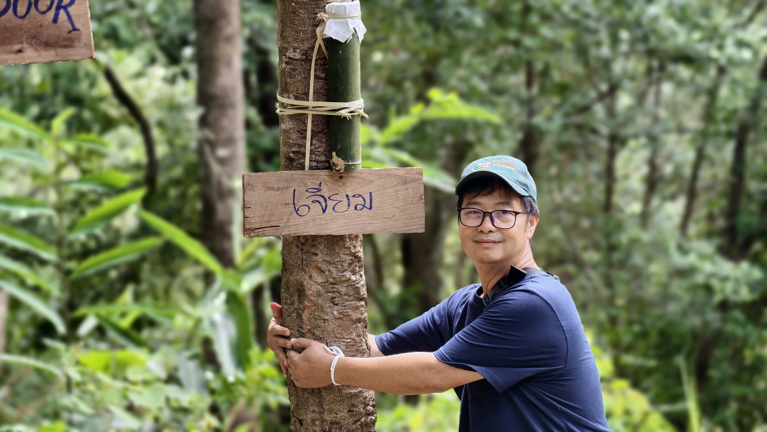 An Indigenous Thai man in glasses and a blue shirt hugging a tree in a lush green forest, with two wooden signs attached to the tree, one above the other, with blue handwriting in Thai.