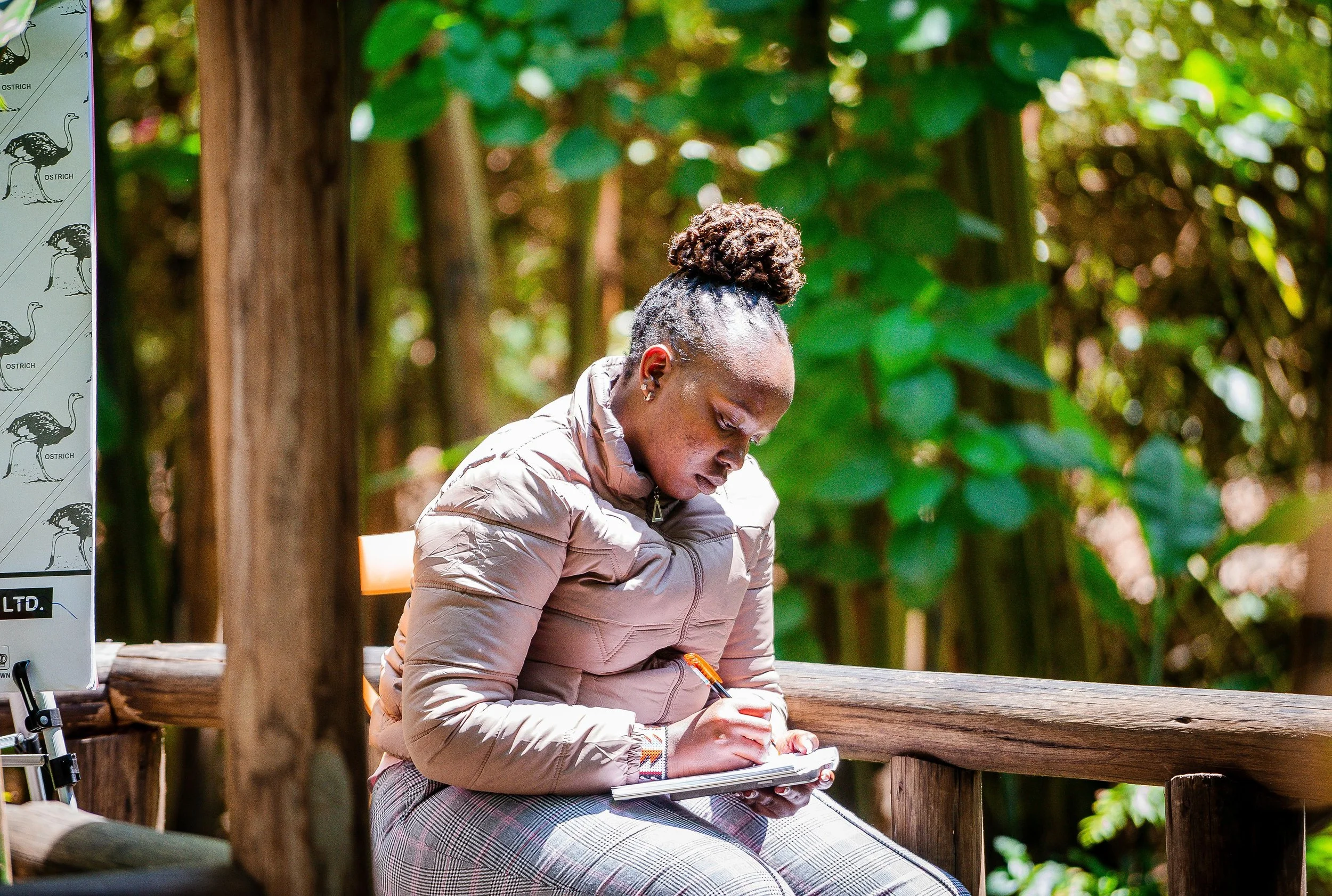 A young Kenyan Indigenous woman sitting on a wooden bench in a green, outdoor environment, writing in a notebook.