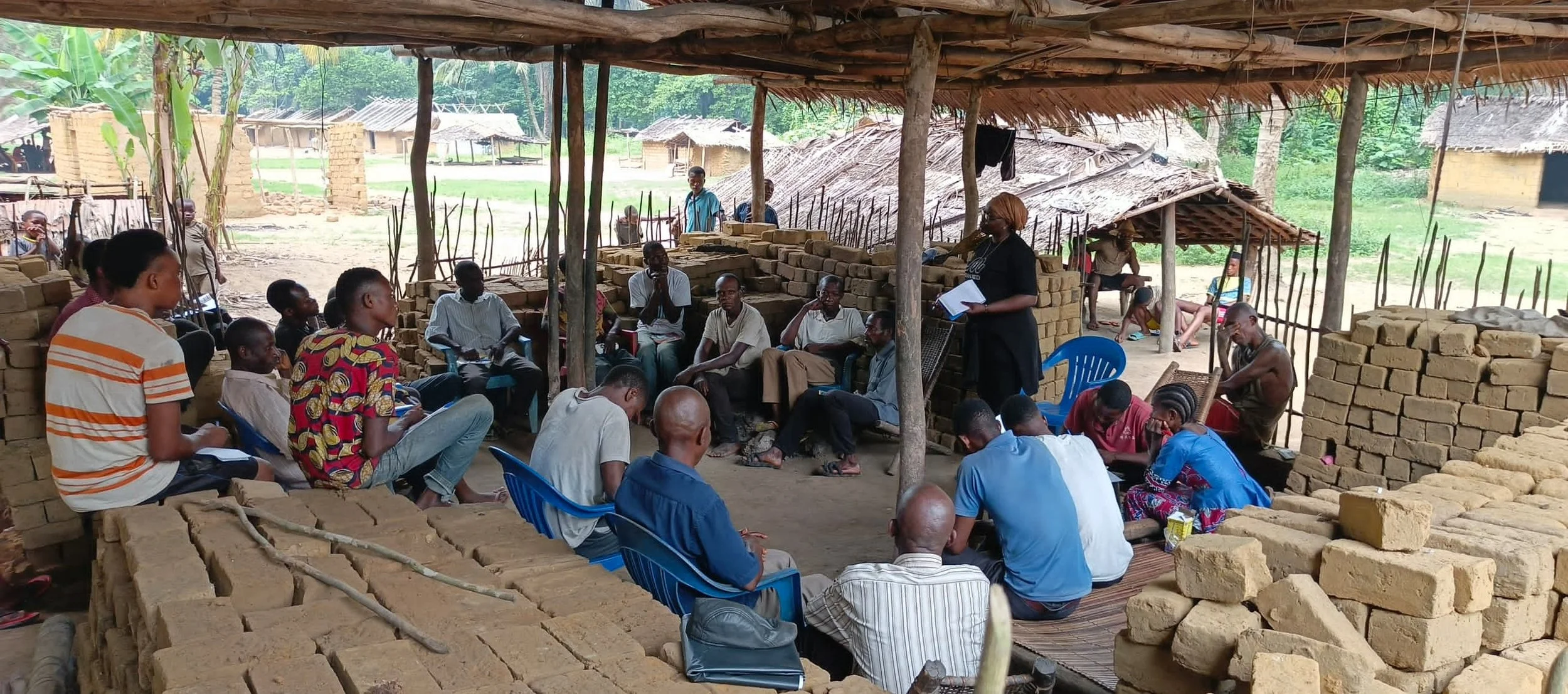 Community meeting taking place in an outdoor shelter made of wood and thatch, with people sitting on chairs and on the ground, surrounded by partially constructed brick walls in a rural area.
