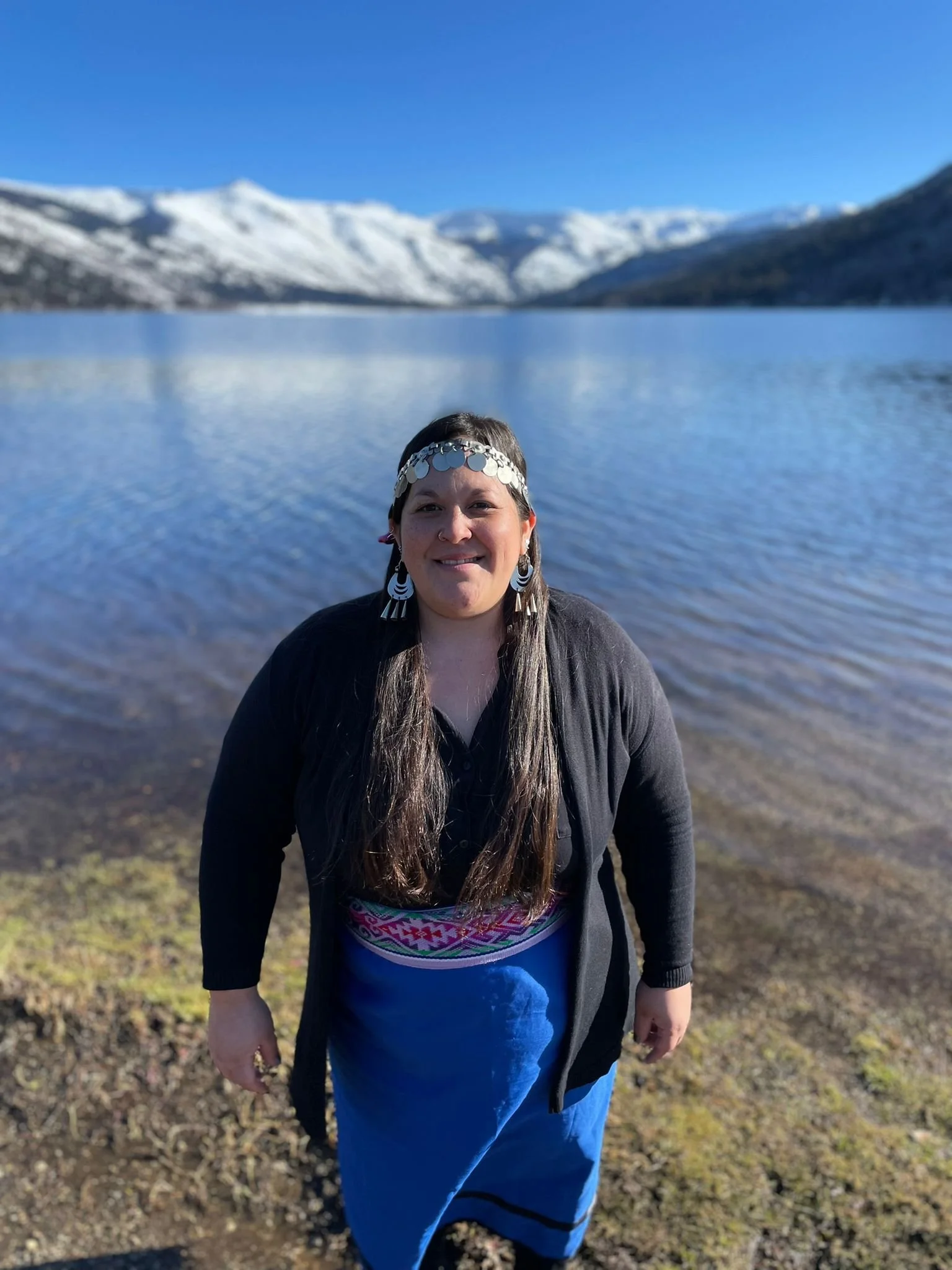 A woman dressed in festival attire, wearing a headpiece with coins, standing outdoors by a lake with snow-capped mountains in the background in the Futa Mawiza Biocultural Territory spanning Argentina and Chile..