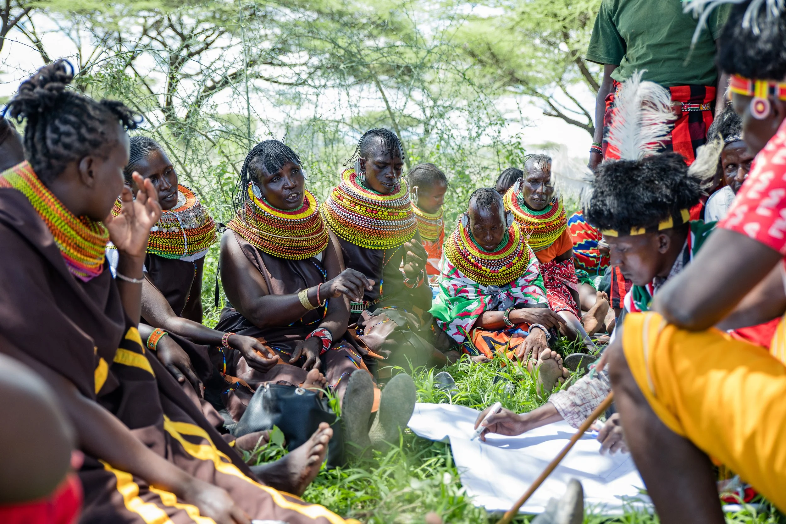 Indigenous African people from Kenya sitting on the grass in traditional attire, engaged in a community gathering or meeting outdoors.