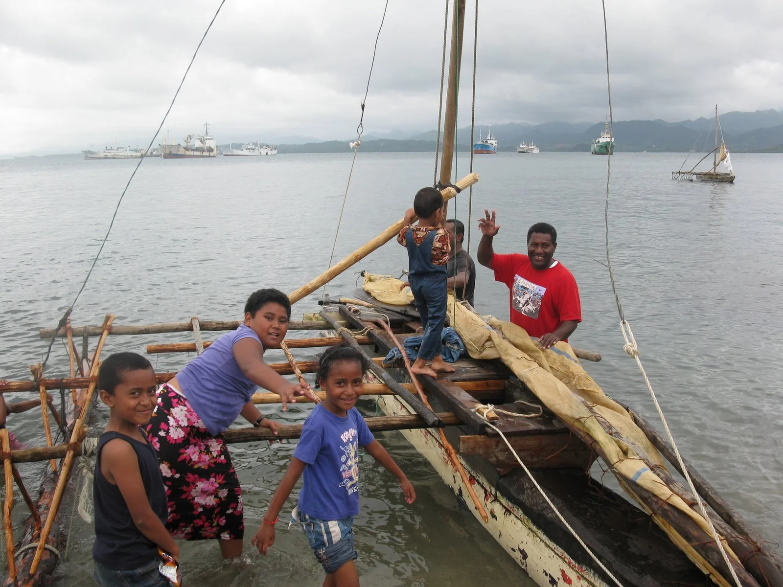 A group of Fijian children and a man standing in shallow water next to a partially constructed wooden boat, with a cloudy sky and multiple ships in the background.