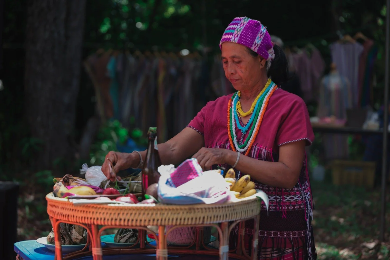 An Indigenous Thai woman wearing traditional clothing and colorful beaded necklaces, standing behind a basket of fresh fruits and vegetables at an outdoor market.