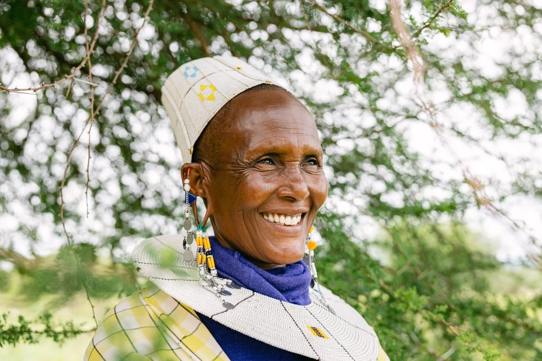 A smiling Maasai woman in Tanzania wearing traditional jewelry and clothing, outdoors with greenery.