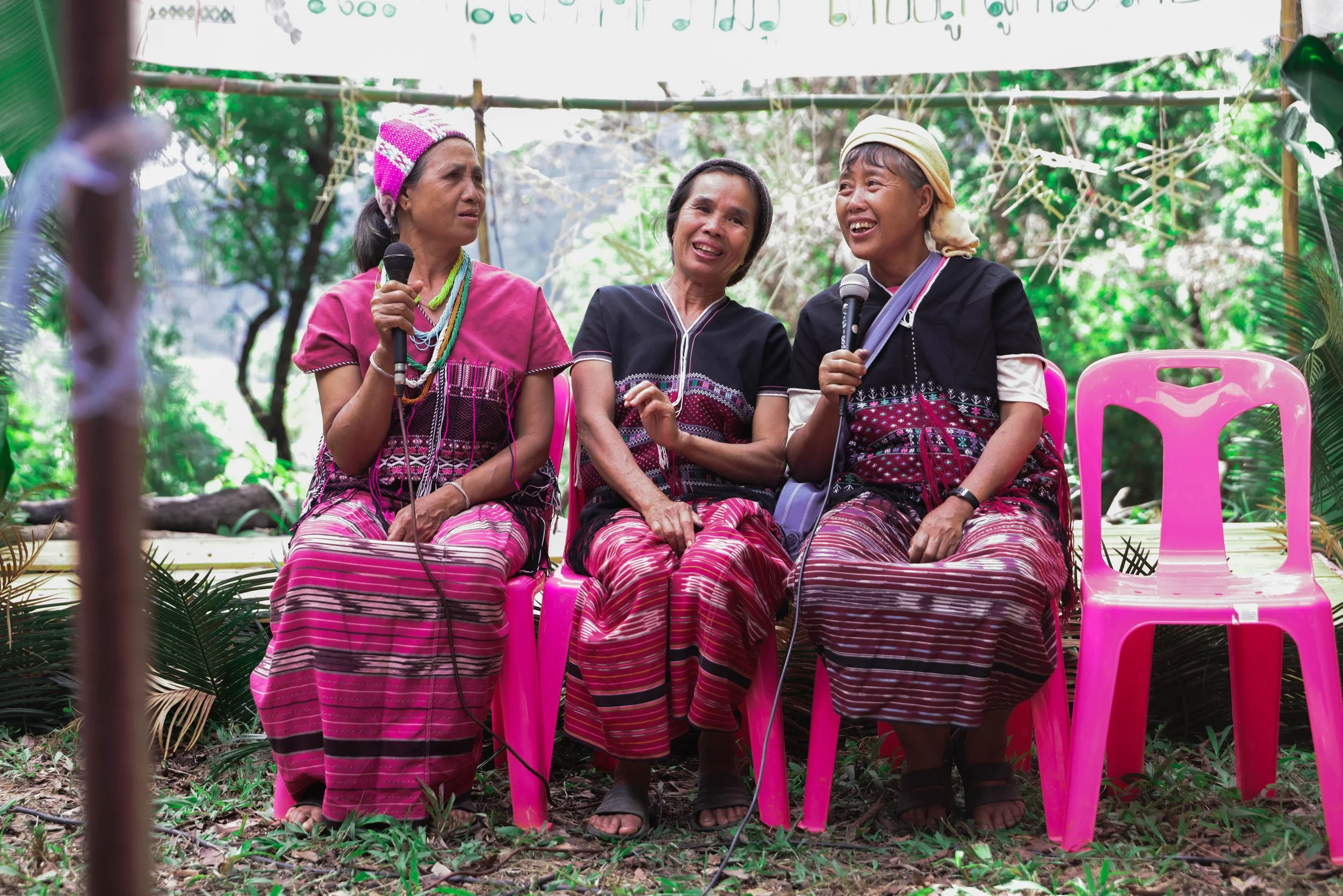 Three Indigenous Thai women in traditional clothing sitting on pink plastic chairs outdoors, holding microphones, surrounded by greenery and decorations.