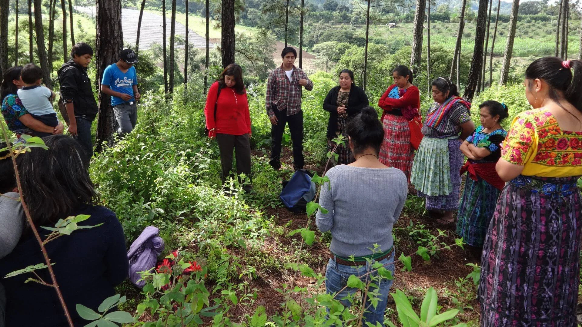 Group of people gathered outdoors in a forest, listening to a woman speaker in the center. The group includes men and women, some wearing traditional clothing, standing and sitting among trees and greenery in Guatemala.