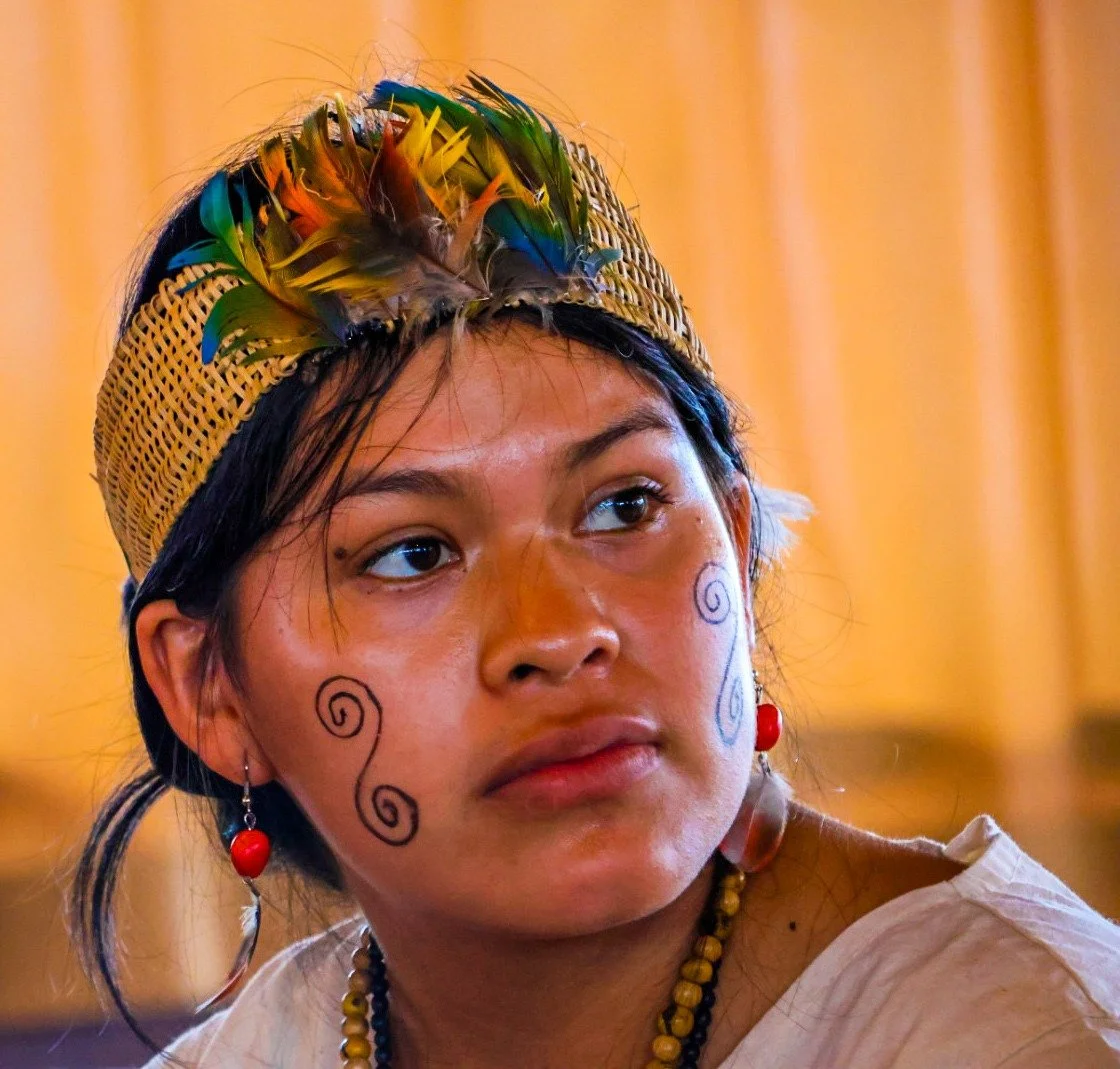Young Indigenous Peruvian woman with face paint, decorated with swirly designs on her cheeks, wearing a woven hat with colorful feathers, earrings, and necklace.