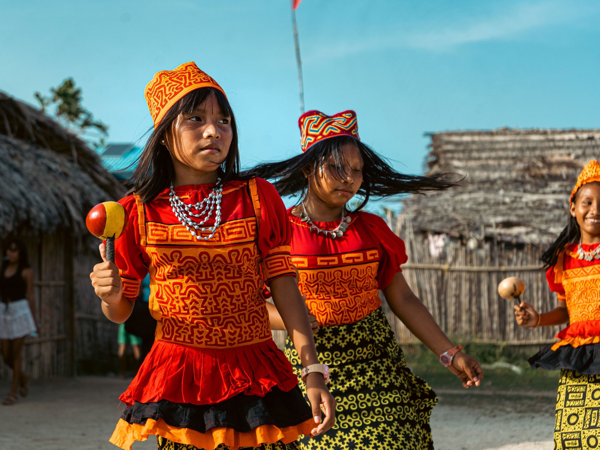 Three young Kuna girls in colorful traditional attire, dancing outdoors with huts in the background, on a sunny day.