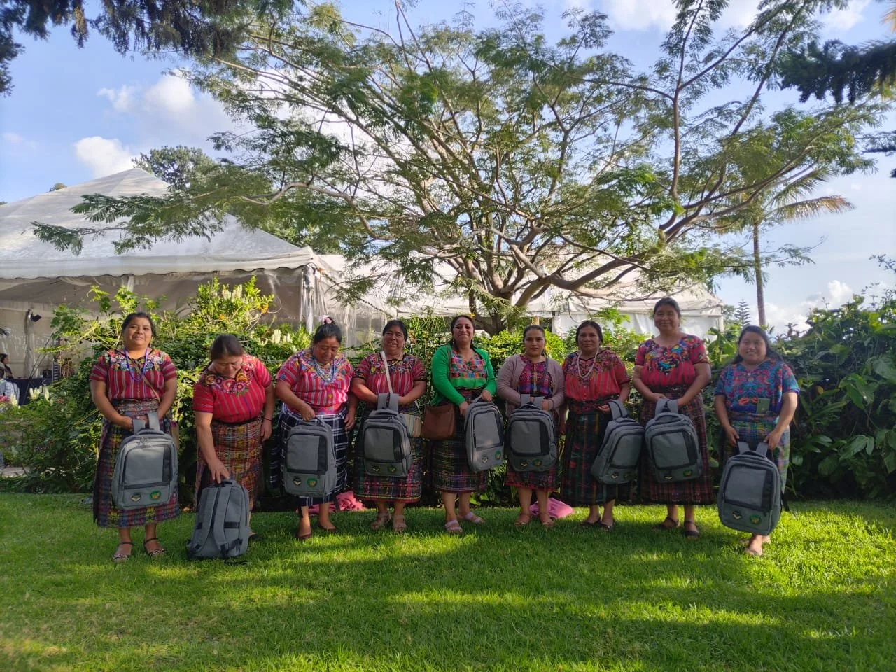 A group of nine Indigenous Guatemalan women dressed in traditional, colorful clothing, standing outdoors on grass, each holding a backpack, with a large tree, a white tent, and a blue sky with clouds in the background.