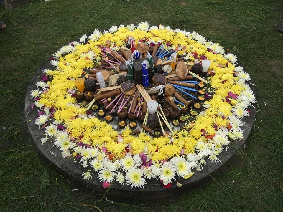 A circular arrangement of sweets and chocolates on a bed of flower petals, chalks, and colored sticks, surrounded by yellow and white flowers on grass as a part of an Indigenous Maya ceremony.