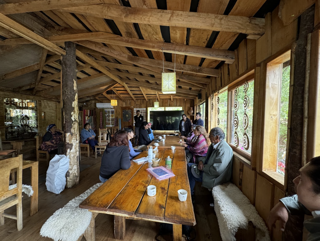 A group of people seated around a long wooden table in a rustic wooden cabin, engaged in a meeting or discussion, with some taking notes and others listening, with large windows on one side showing greenery outside in the Futa Mawiza Territory.