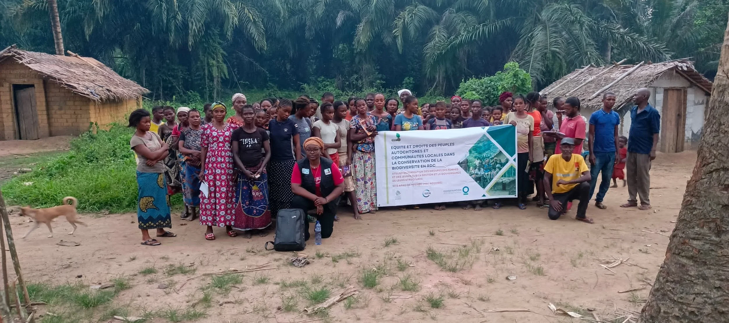 Group of villagers and officials holding a banner about indigenous peoples' rights and biodiversity conservation in a rural area with palm trees and traditional huts in the DRC.