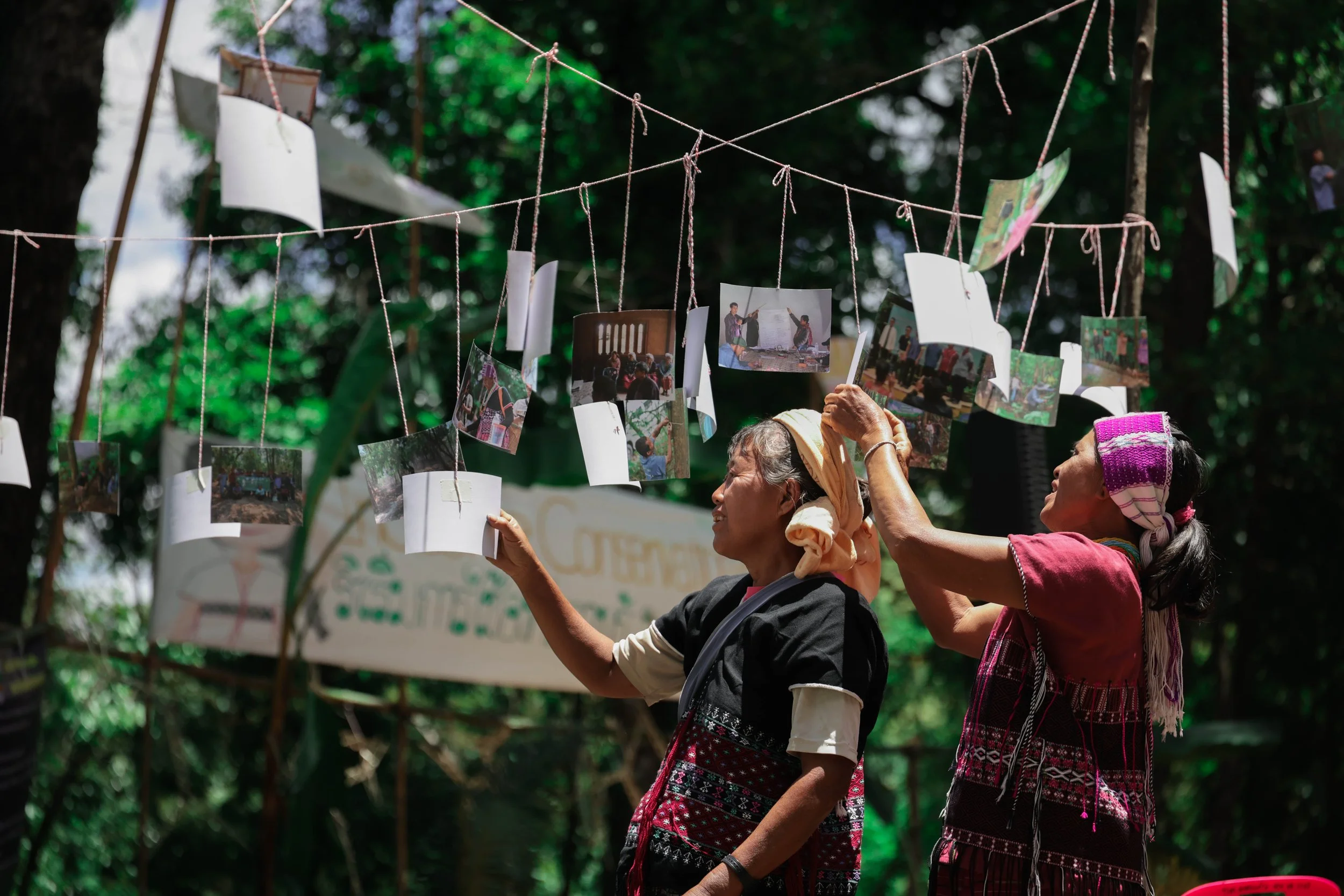 Two Indigenous Thai women in traditional clothing hanging photographs and papers on a string outdoors surrounded by greenery.