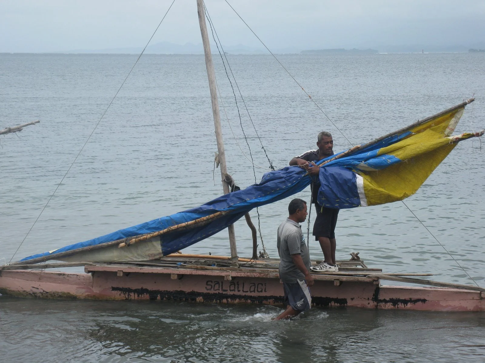 Two men adjusting a small sailboat on calm water with cloudy sky in the background in Fiji.