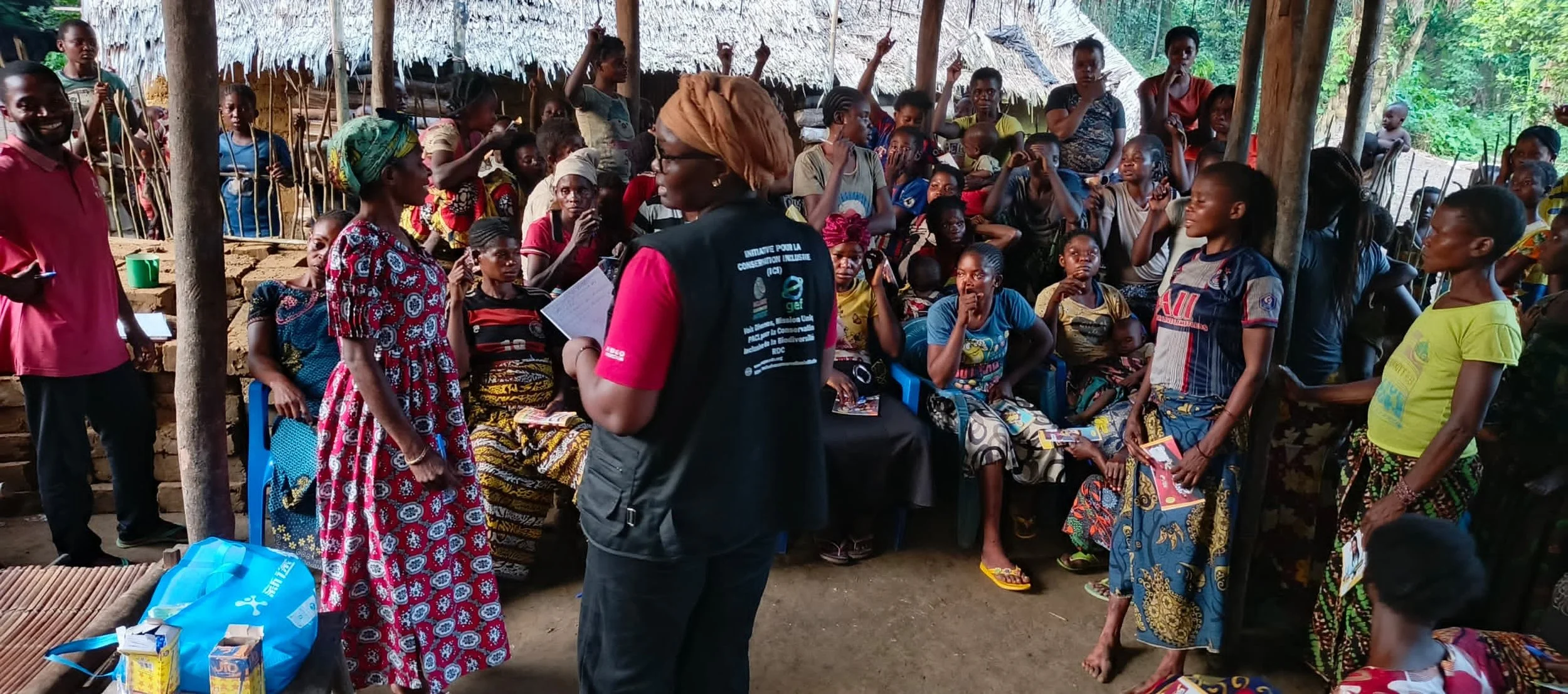 A large group of women and children gathered in a rustic open-air setting in the DRC, some sitting and some standing, engaged in a community meeting or event. Two women are standing and speaking, while others are listening or taking photos.