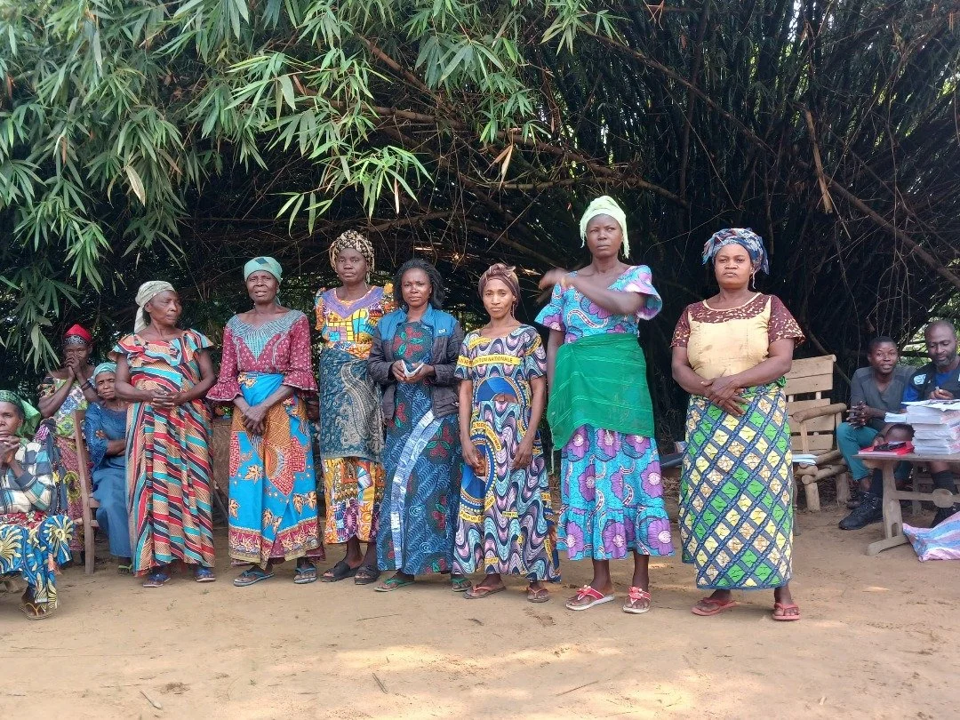 Group of Indigenous women standing outdoors in the DRC, wearing colorful traditional dresses with a large bush or tree in the background.