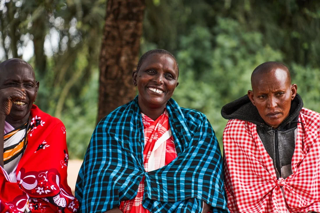 Three Kenyan women sitting outdoors under a tree, smiling and wearing colorful traditional Maasai clothing.