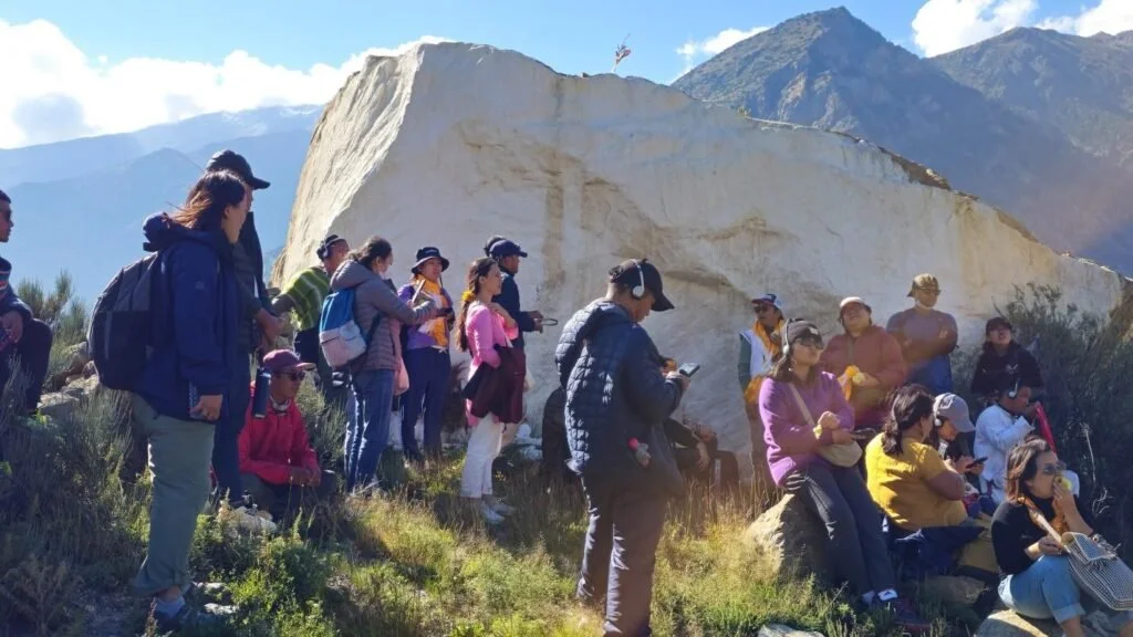 Group of people gathered outdoors near a large white rock formation, with mountains in the background, some standing and some sitting, some using phones or listening to a guide, under a clear sky in the Annapurna Area near the Himalayas.